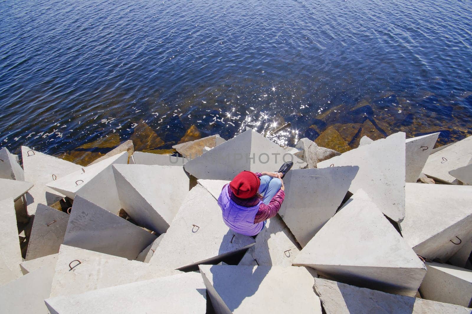 A young woman in a hat on a sunny spring day sits on the breakwaters and admires the sea. Girl in warm clothes on the breakwater