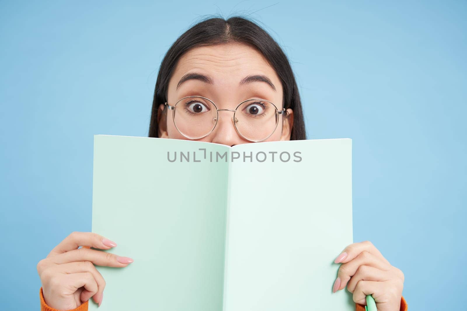 Cute asian woman in glasses, hides her face behind notebook and smiles with eyes, curious gaze, stands over blue background. Students and education concept.