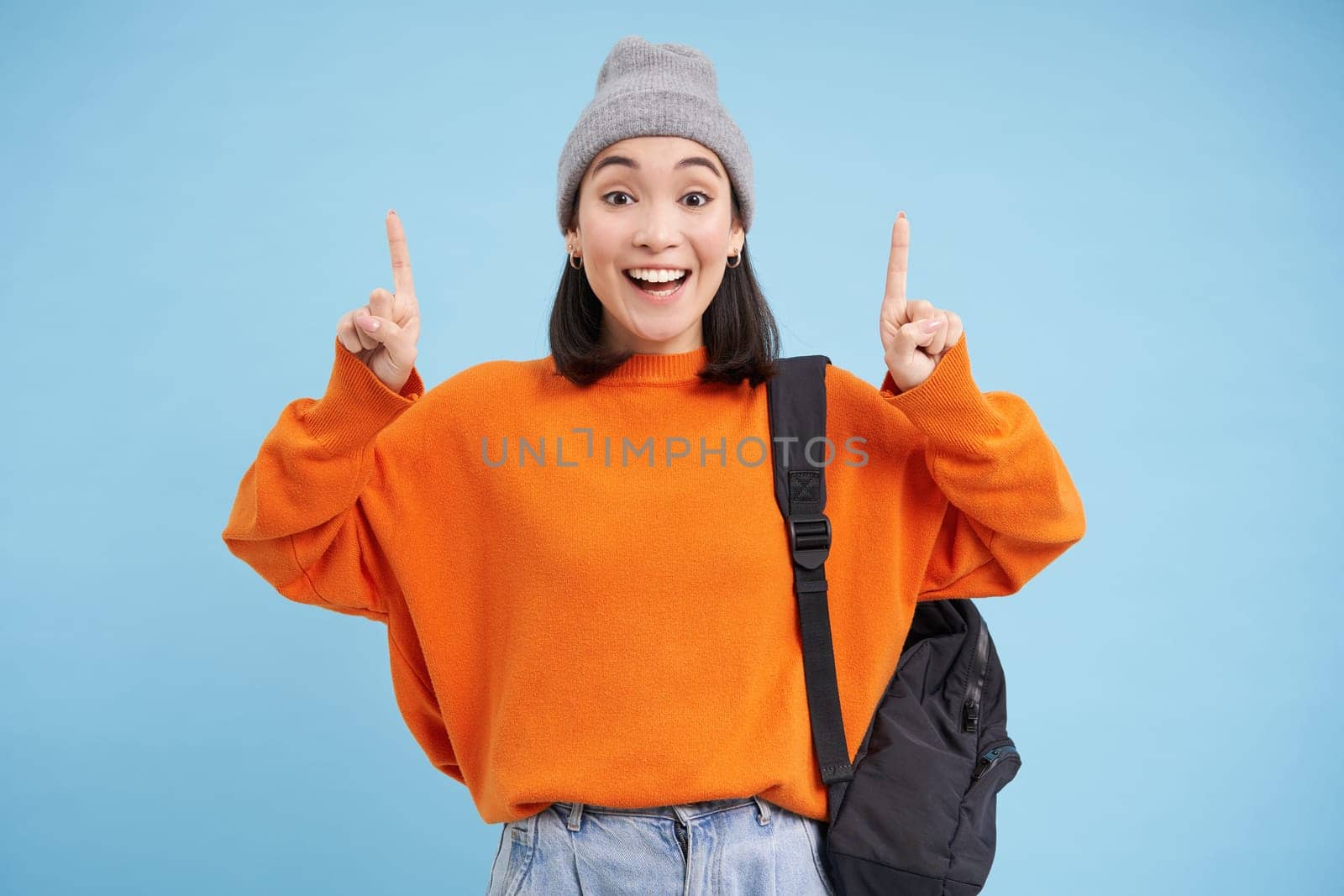Happy korean girl points fingers up, shows advertisement, invites to click banner on top, stands over blue background.