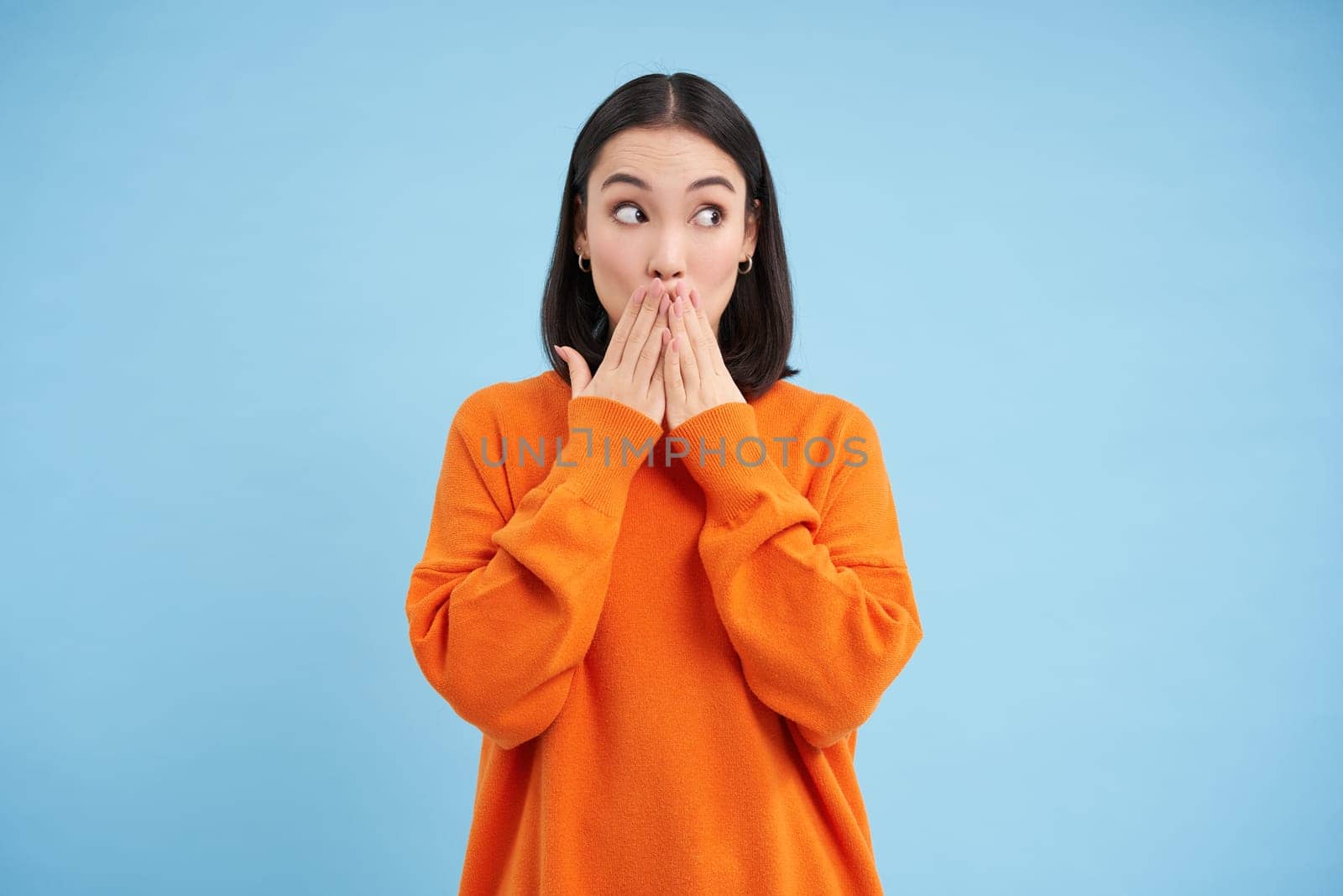 Image of coquettish korean woman, looks aside at smth interesting, holds hands on mouth, amazed by news or gossips, stands over blue background.