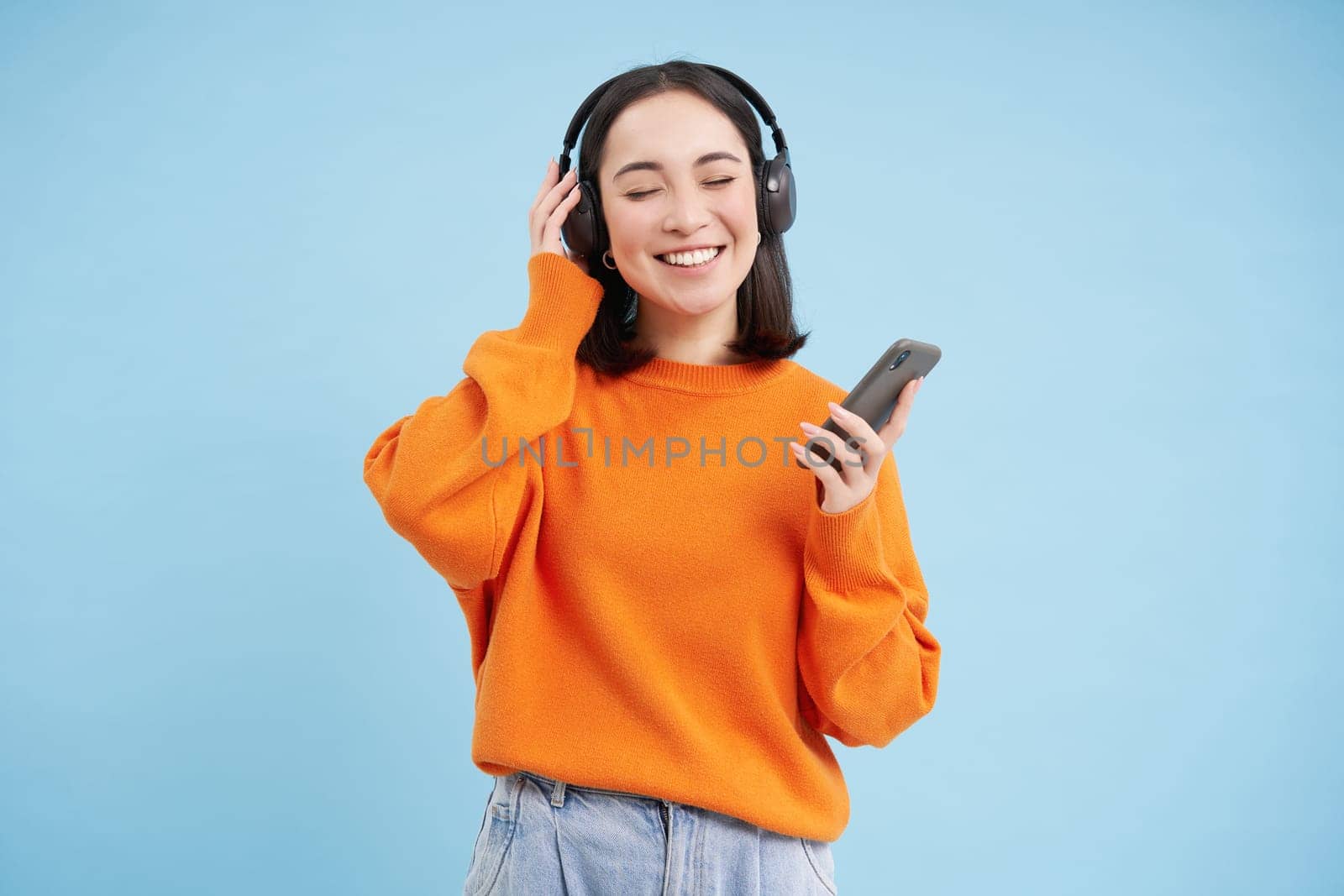 Beautiful young woman in headphones with mobile phone, listens to music in earphones and smiles, blue background.