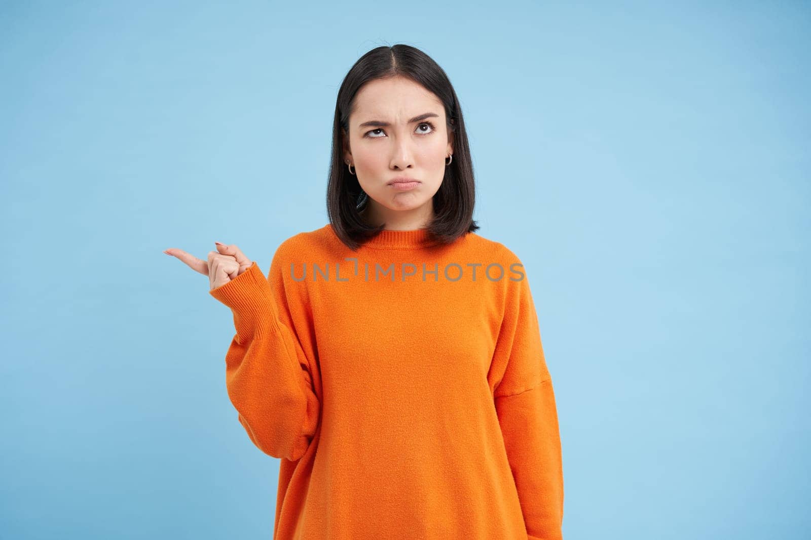 Confused korean girl cant understand something, points left with clueless face, thinking, standing in orange shirt against blue background by Benzoix