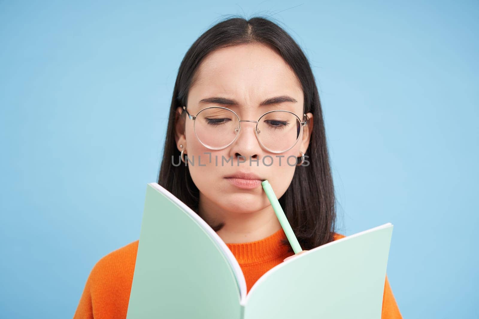 Portrait of thinking asian woman in glasses, solves task in notebook, writes down exercise, student learning, doing homework, blue background by Benzoix