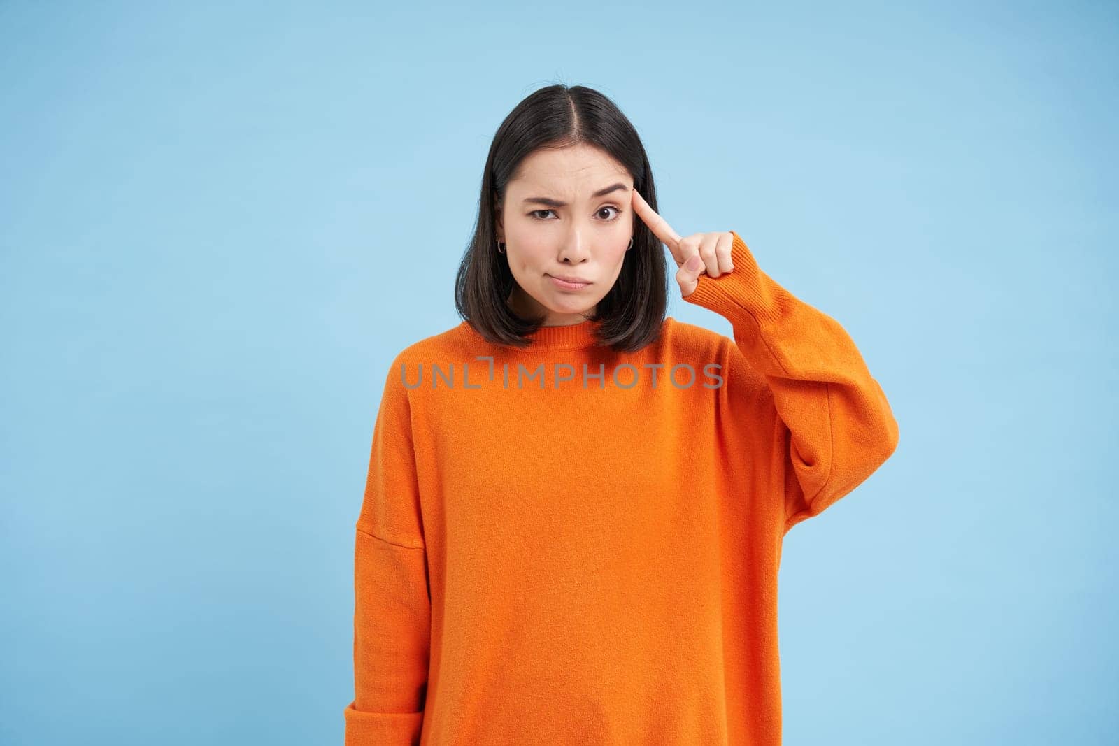 Are you crazy. Young skeptical asian woman holds finger near head, disappointed by someones behaviour, stands over blue background.