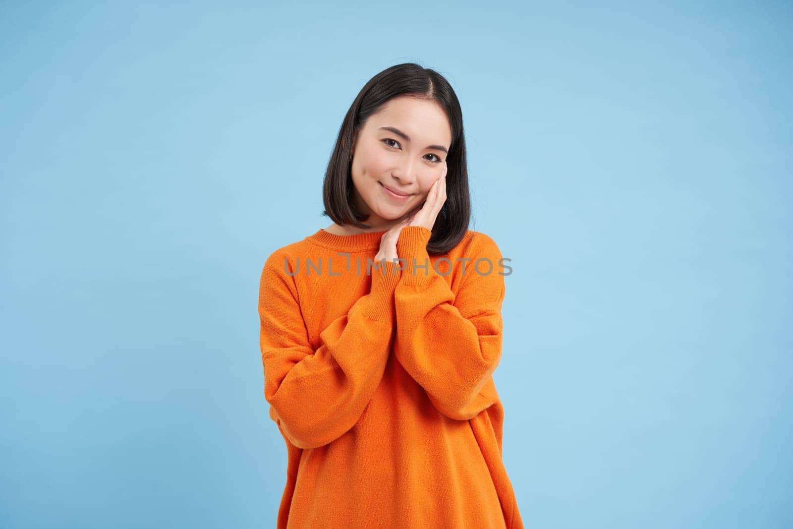 Beauty and wellbeing. Young brunette woman laughing and smiling, touching her natural healthy face, standing over blue studio background.