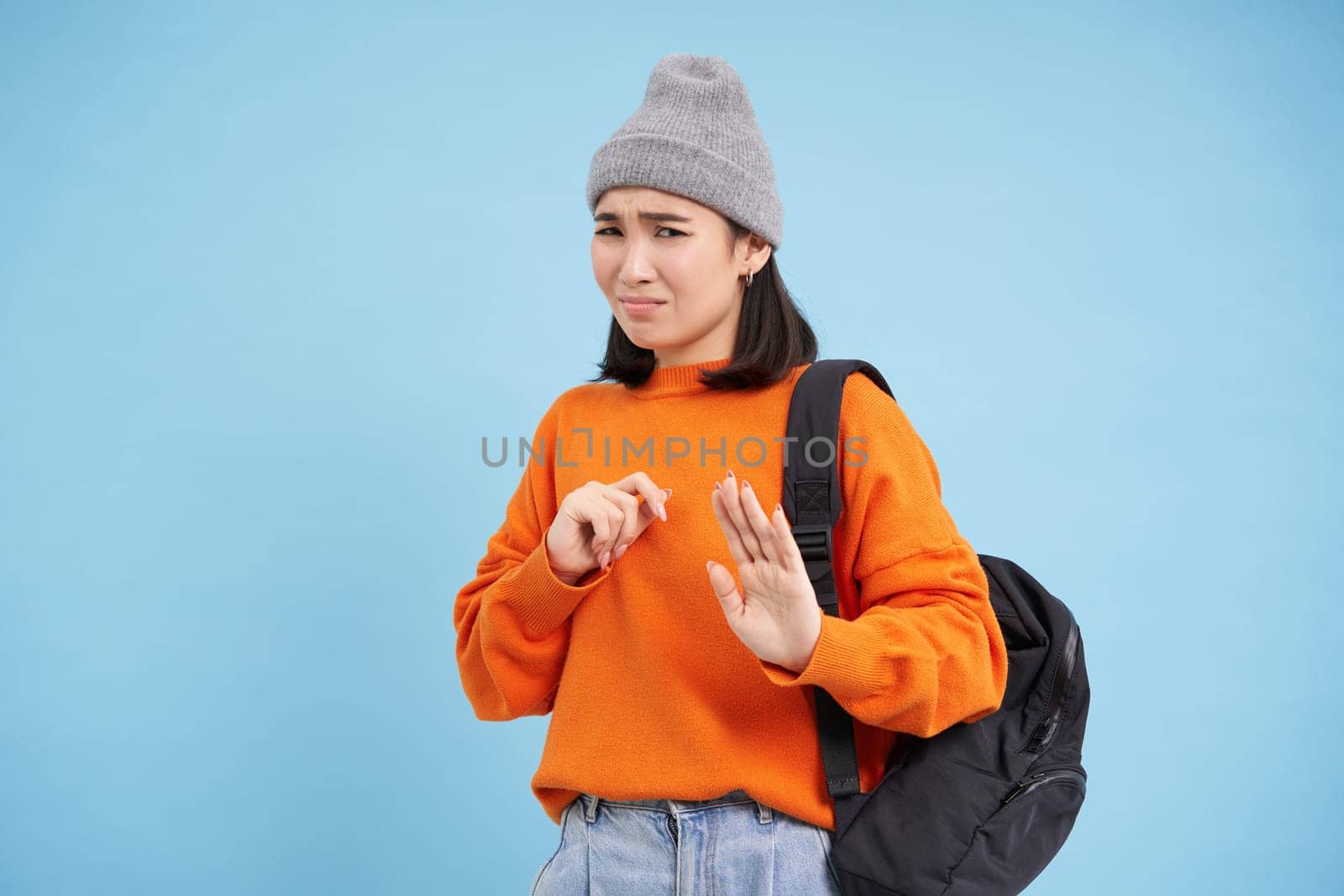 Asian girl grimaces, shows block hands gesture, stop sign, asks to stay back, rejects smth disgusting, stands over blue background.