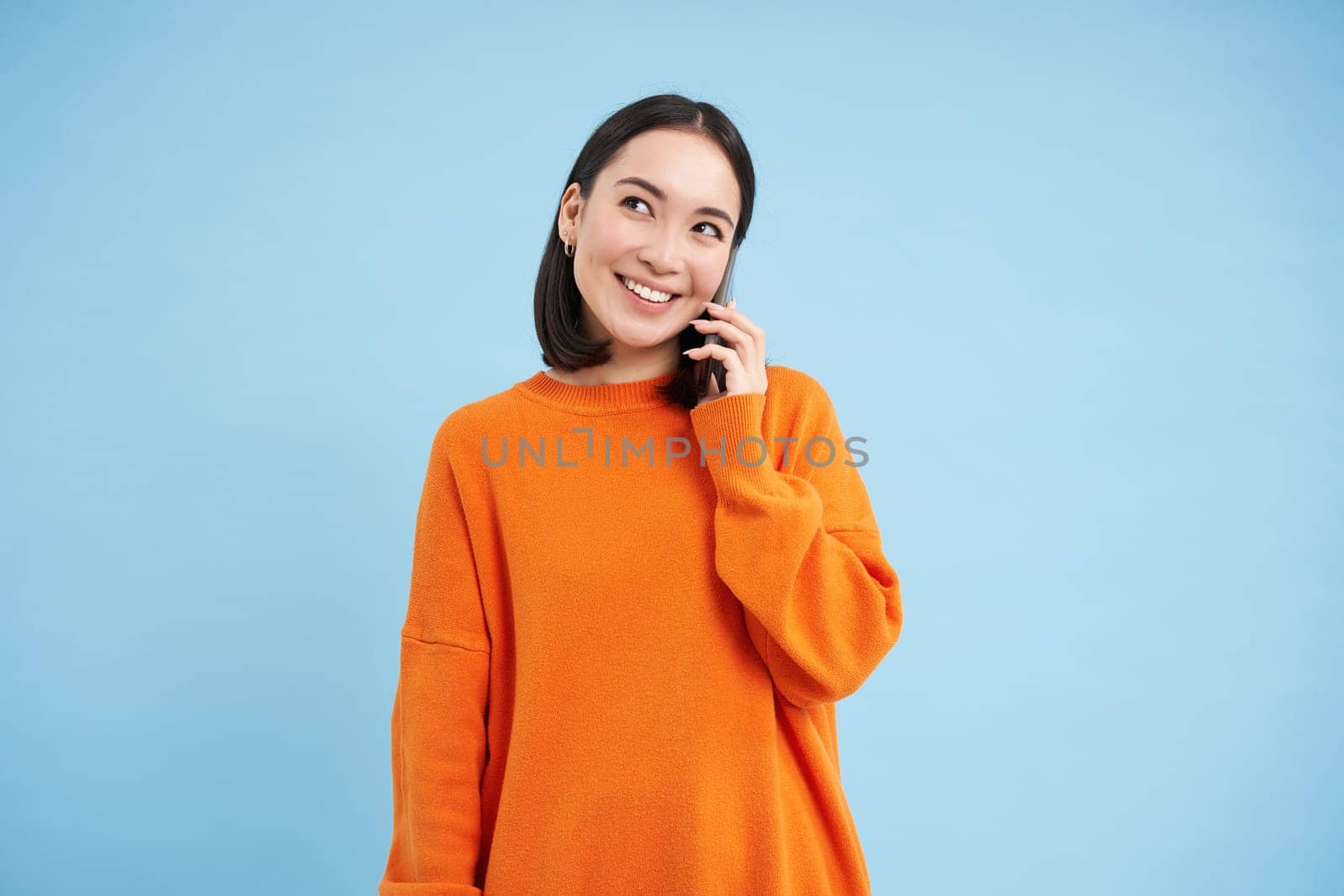 Portrait of young candid woman, asian girl talking on cellphone, holds mobile phone and speaks, stands over blue background.