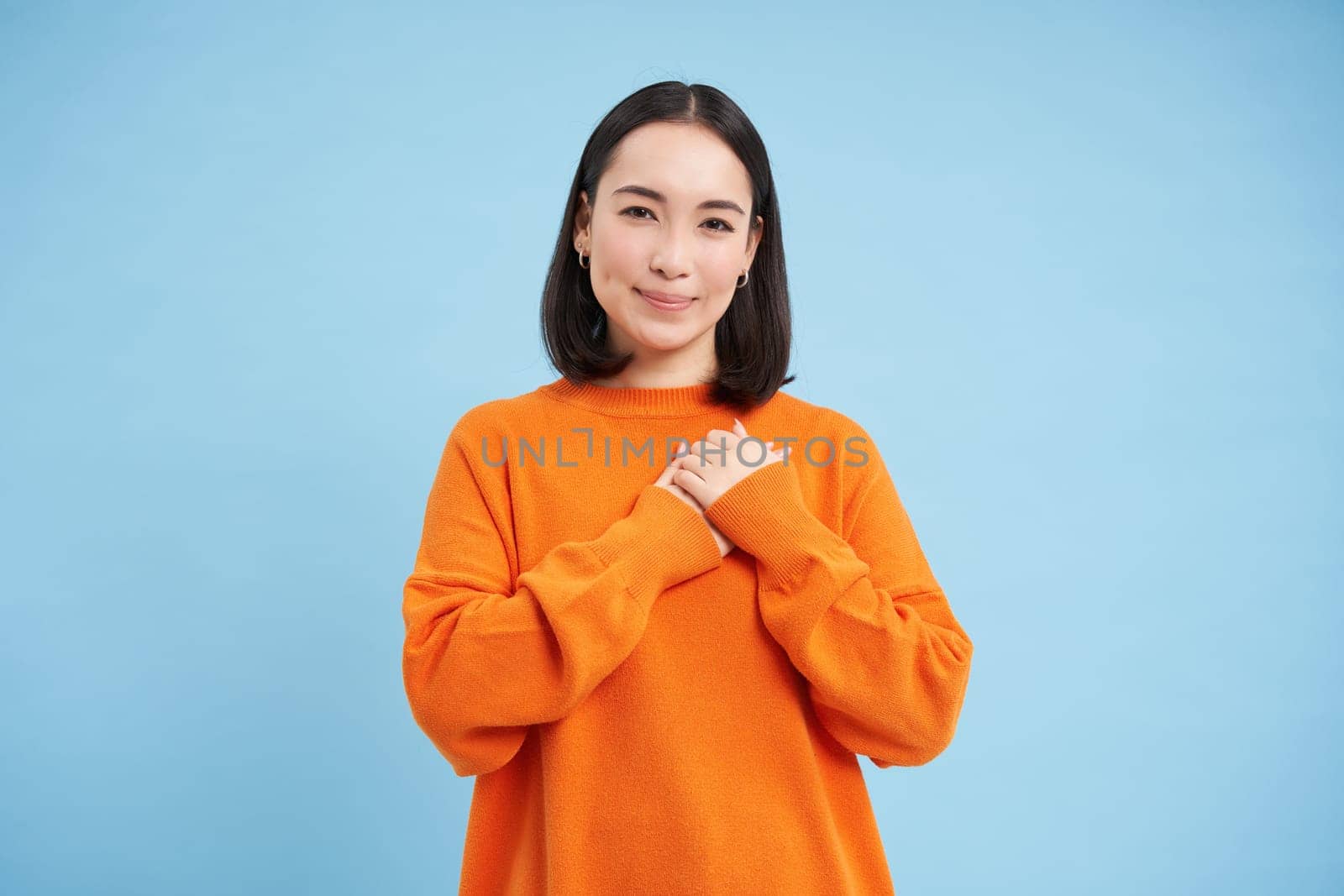 Care and love. Asian woman holds hands on heart and smiles, looks with tenderness and warmth at camera, stands over blue background.