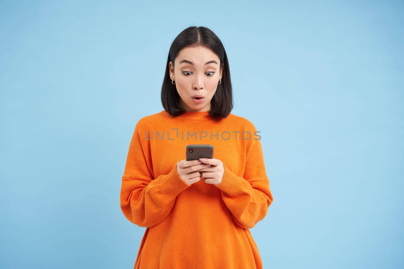 Young woman looks amazed at her cellphone, reads exciting news, found good deal online on mobile application, standing over blue background.