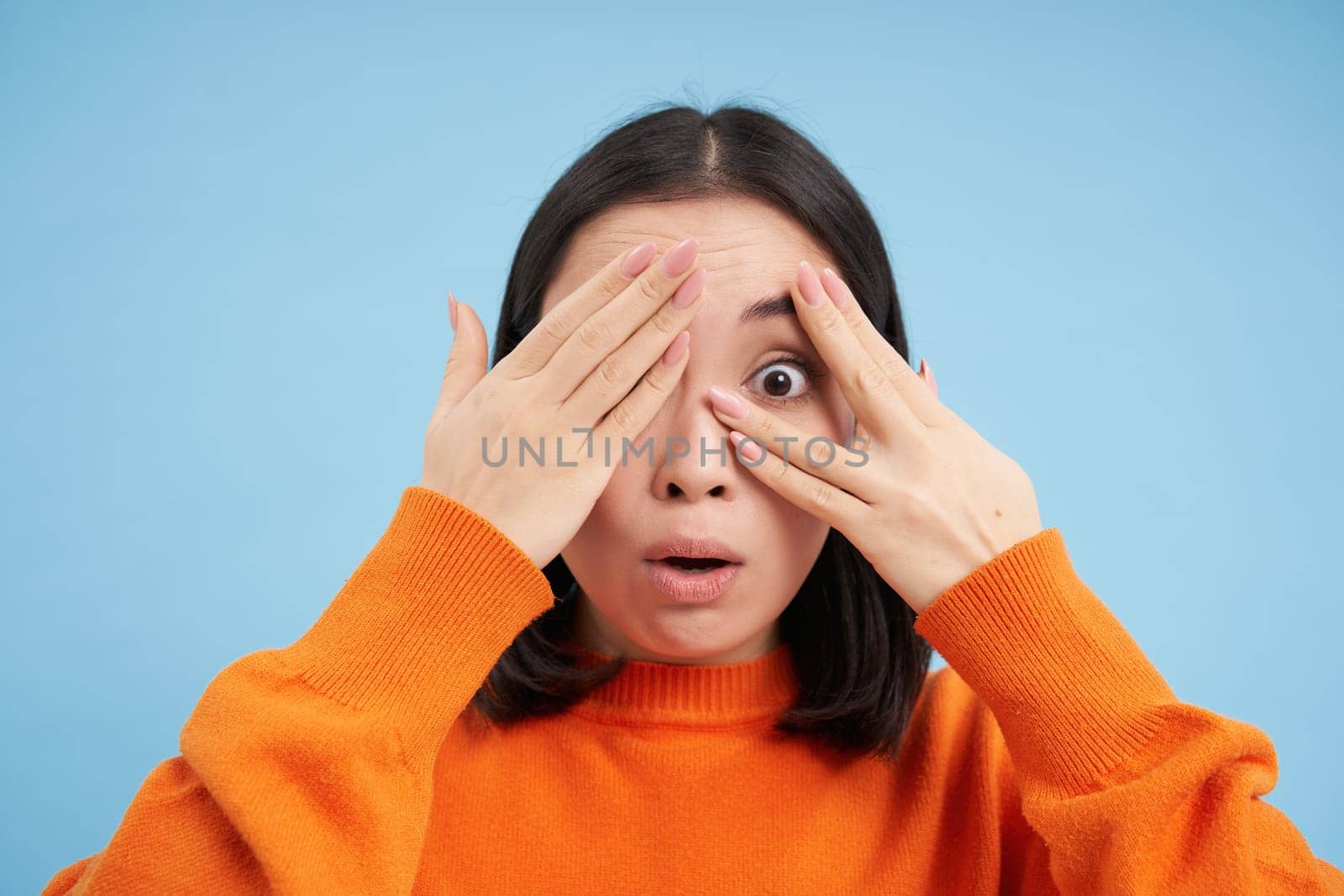 Close up portrait of korean girl waits for surprise, shuts her eyes and peeks through fingers with excited smile, stands over blue background.