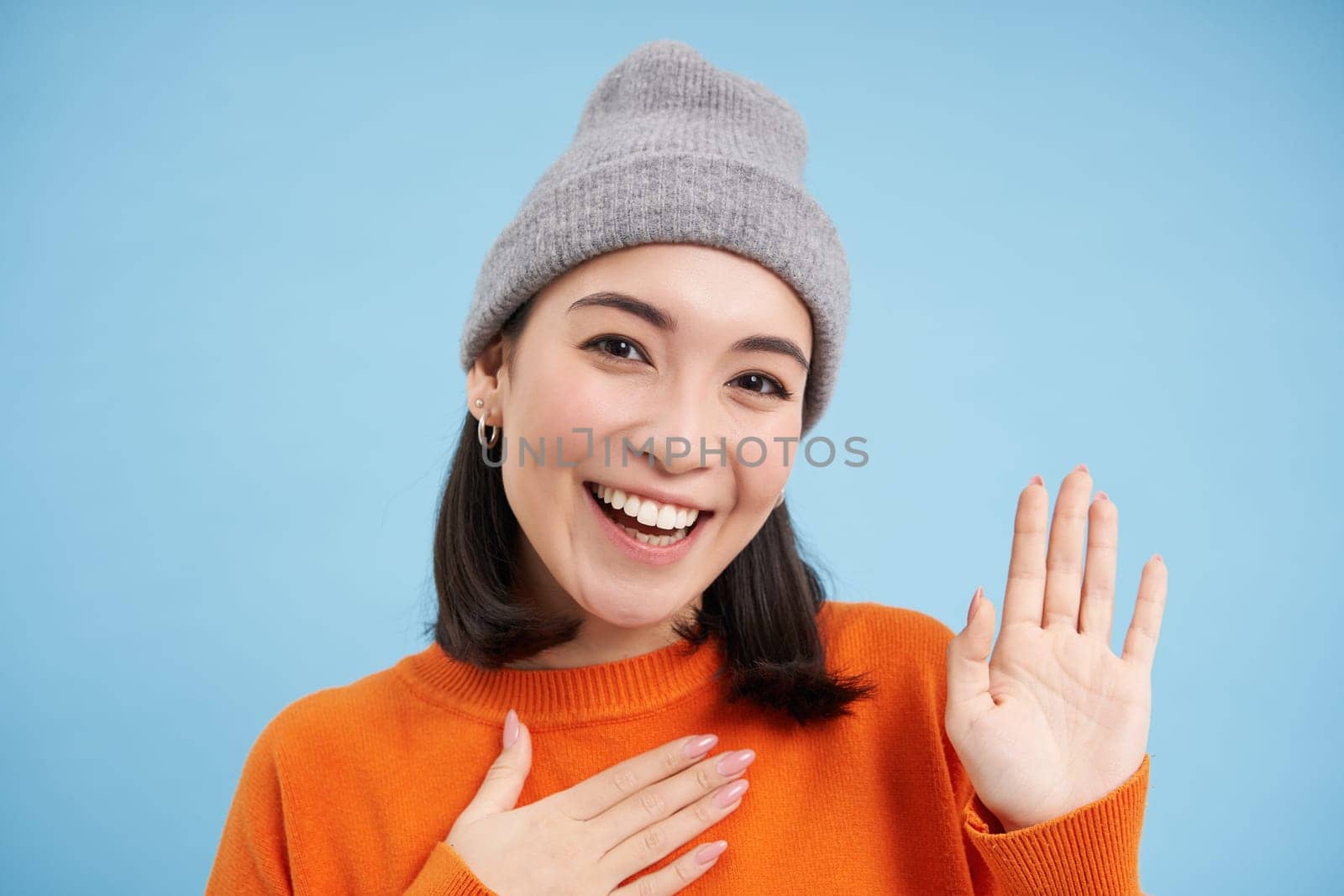 Close up of cute korean girl in beanie, raises one hand and introduces herself, greets you, smiles and looks friendly, blue background.