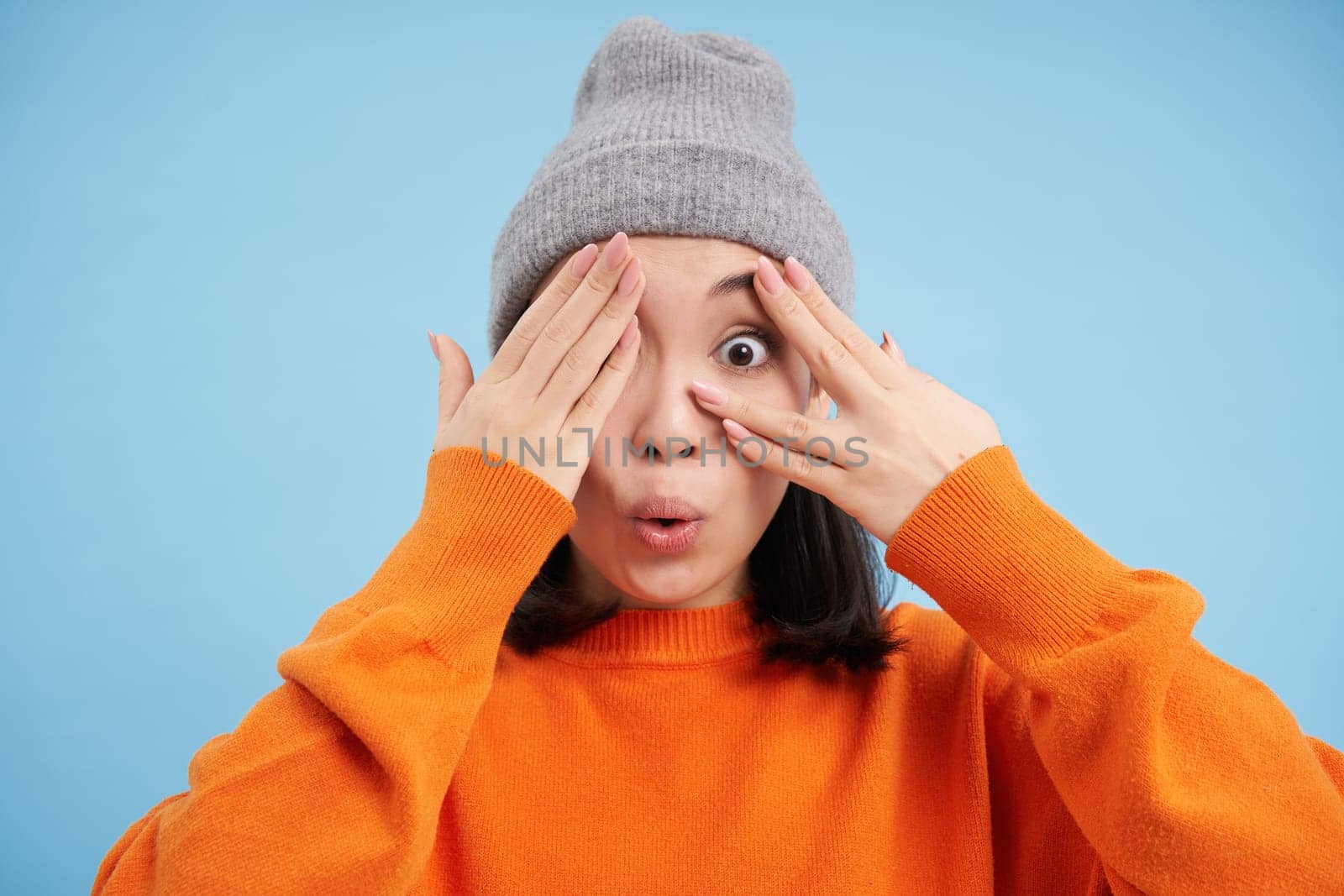 Surprised korean girl in warm hat, looks amazed at camera, expresses interest and amazement, stands in orange sweatshirt over blue background.