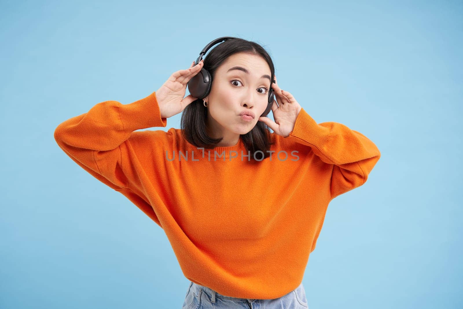 Beautiful korean woman in headphones, dancing and listening music in earphones, standing over blue background.