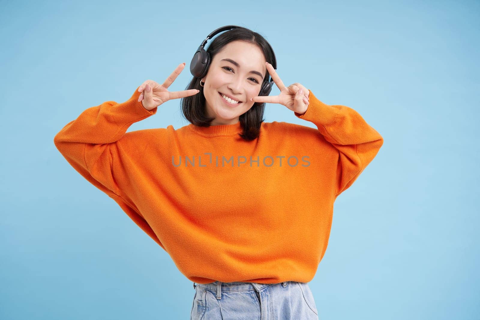 Beautiful korean woman in headphones, dancing and listening music in earphones, standing over blue background by Benzoix