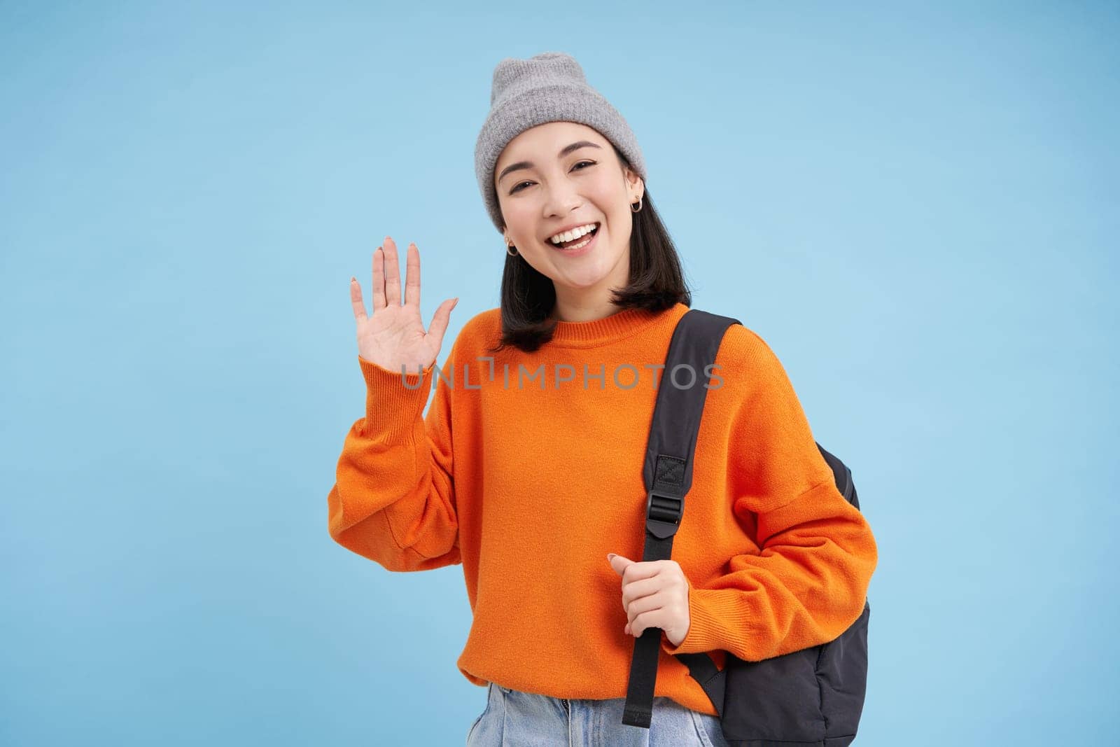 Friendly girl says hello, holds backpack and waves hand, greets you and laughs, stands over blue background.