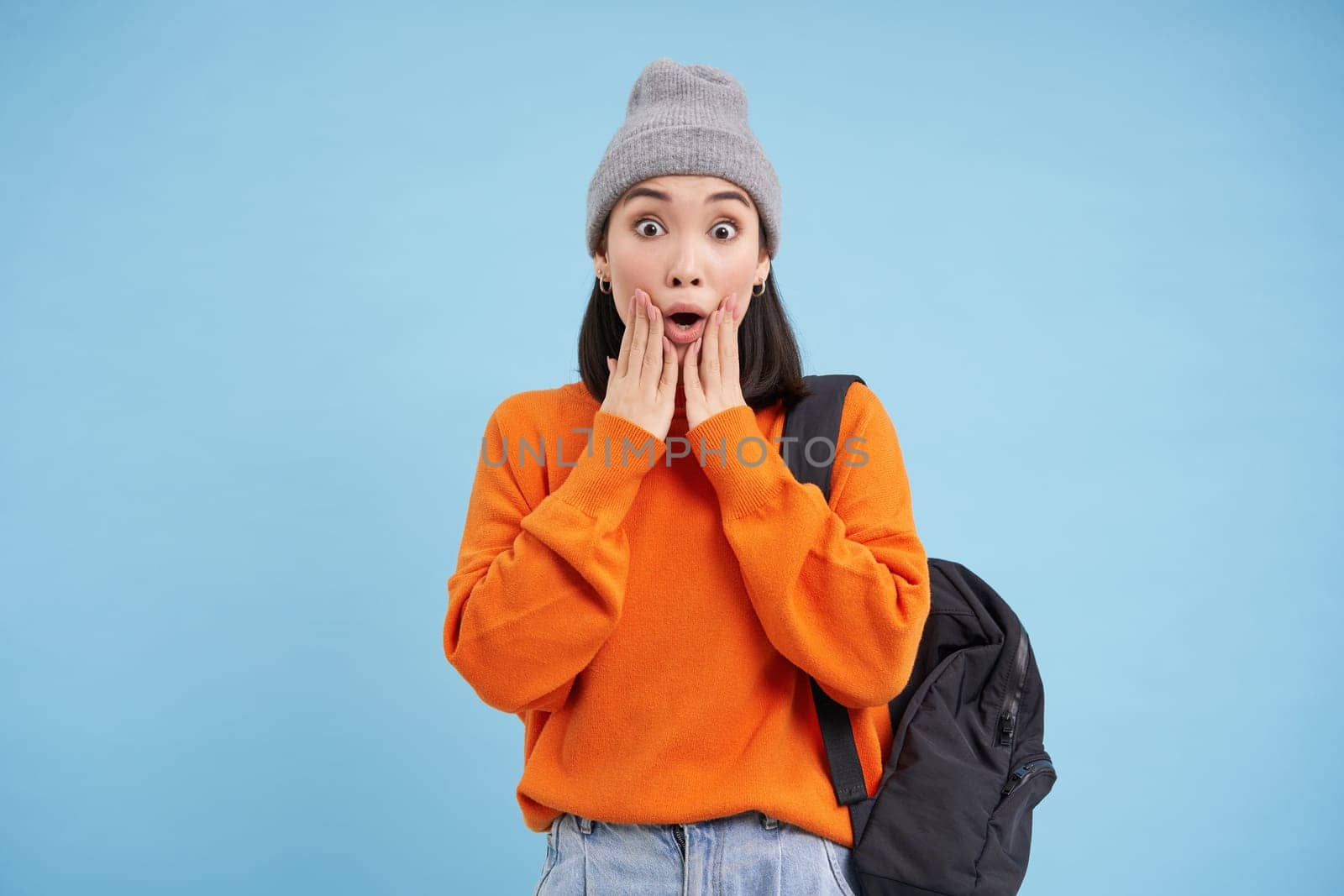 Asian girl in street outfit looks shocked at camera, drops jaw and screams scared, holds backpack, blue background.