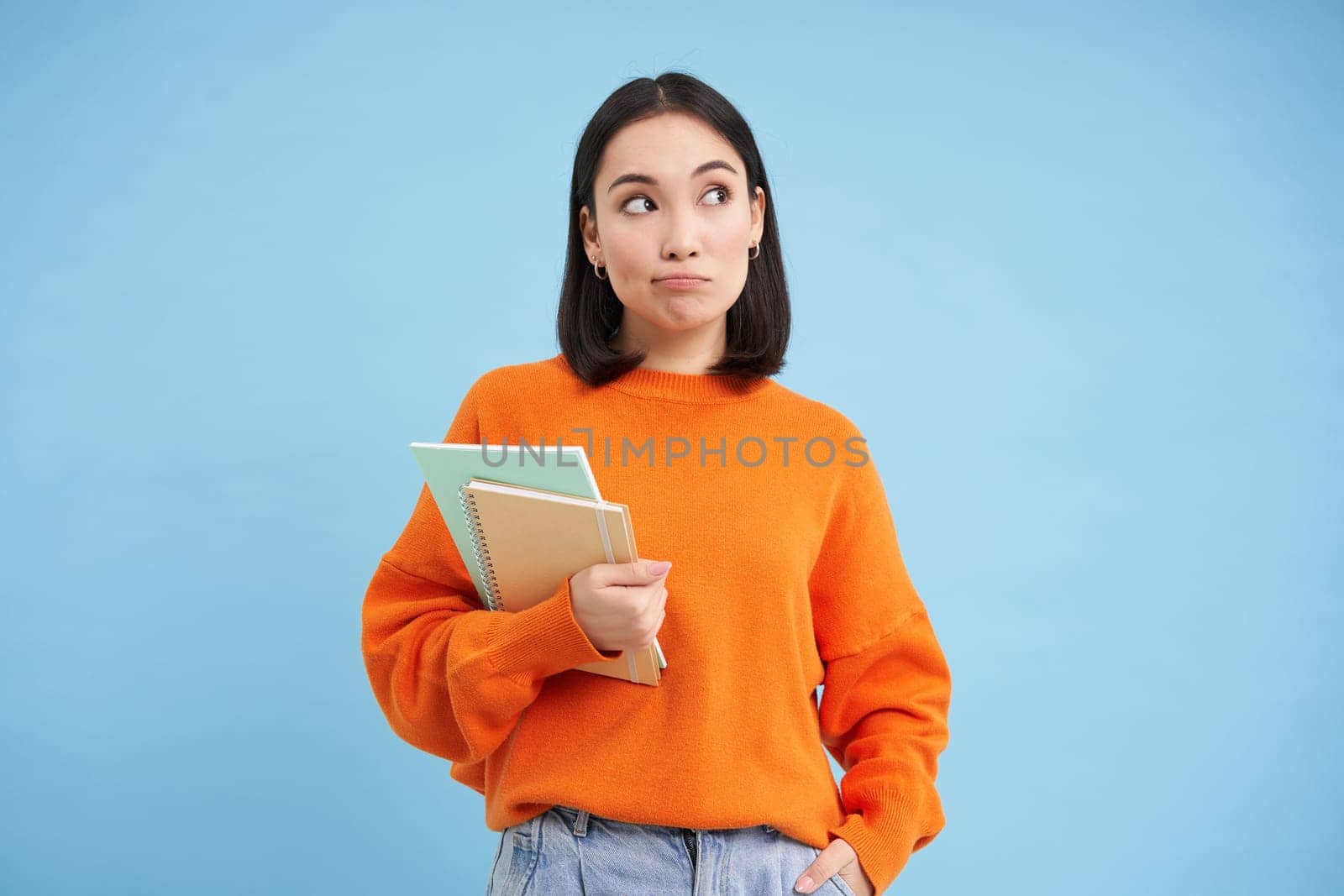 Thinking girl, student with notebooks, stands over blue background thoughtful, making decision.