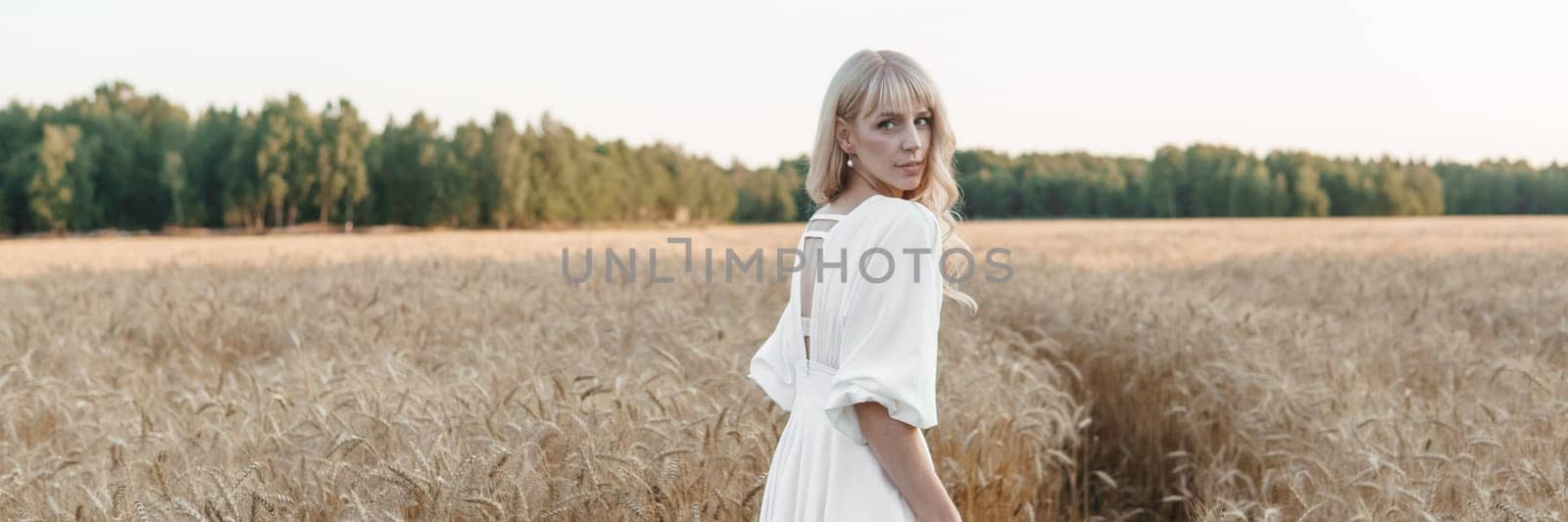 A blonde woman in a long white dress walks in a wheat field. The concept of a wedding and walking in nature