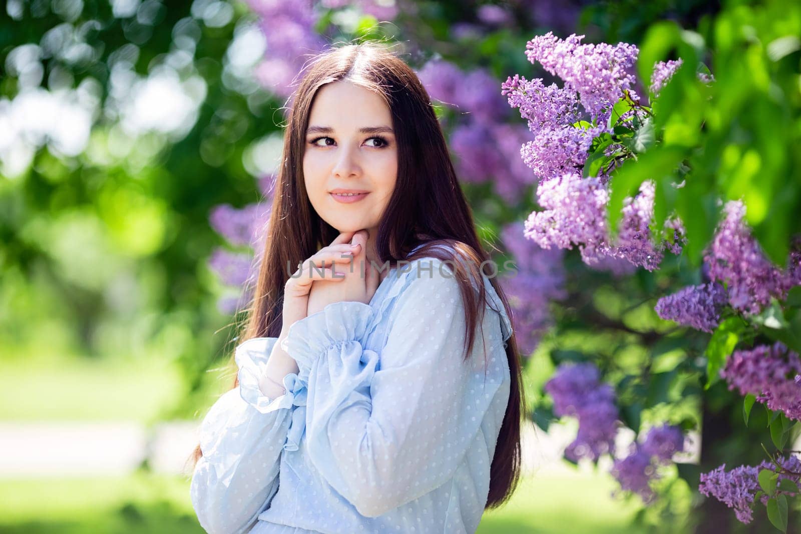 A beautiful girl with long dark hair stands next to a flowering lilac bush in the park. Close up. Copy space