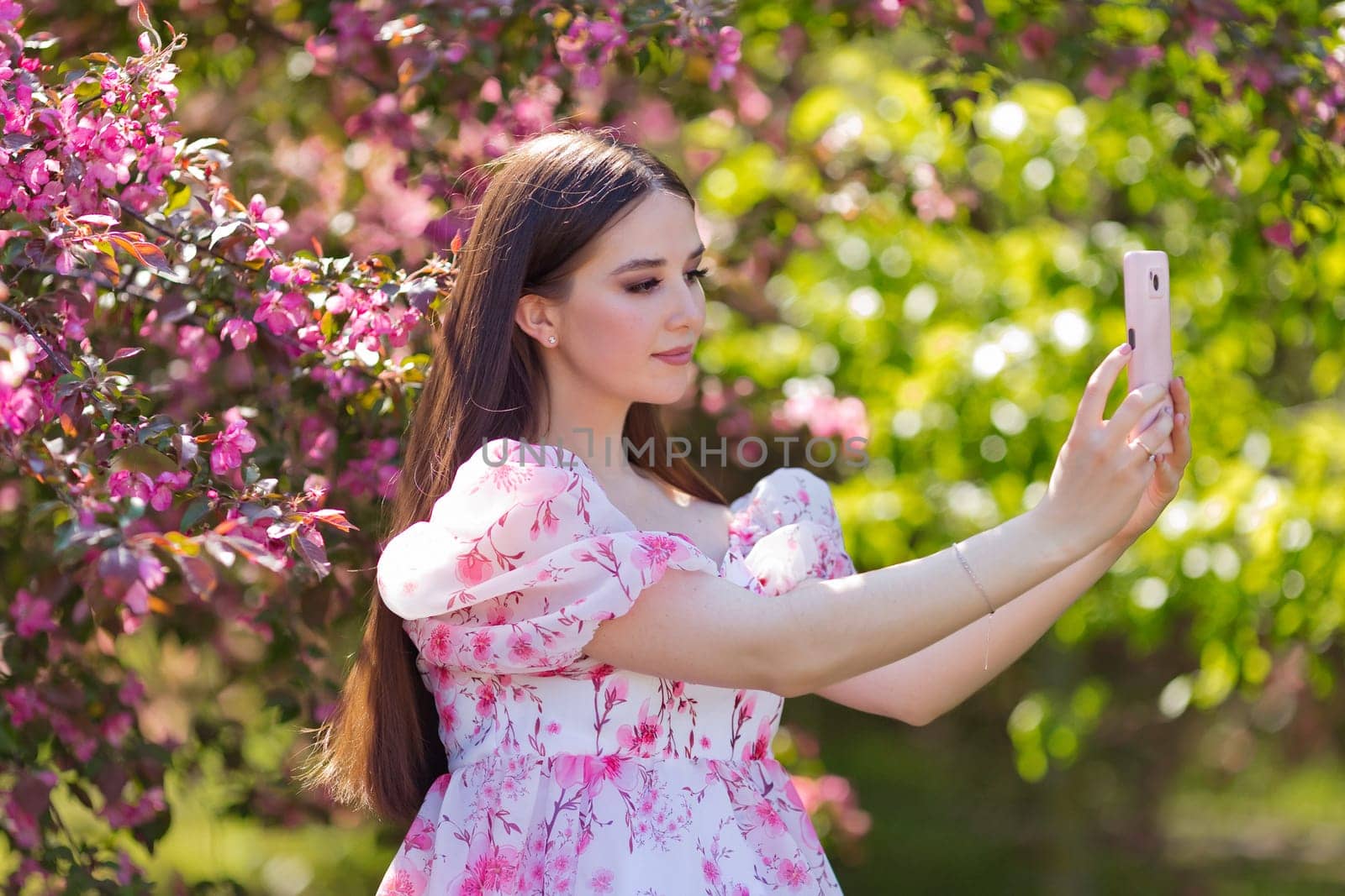 A pretty brunette girl with long hair, makes selfie on smartphone, is standing in a pink blooming apple garden, on a sunny day. Close up