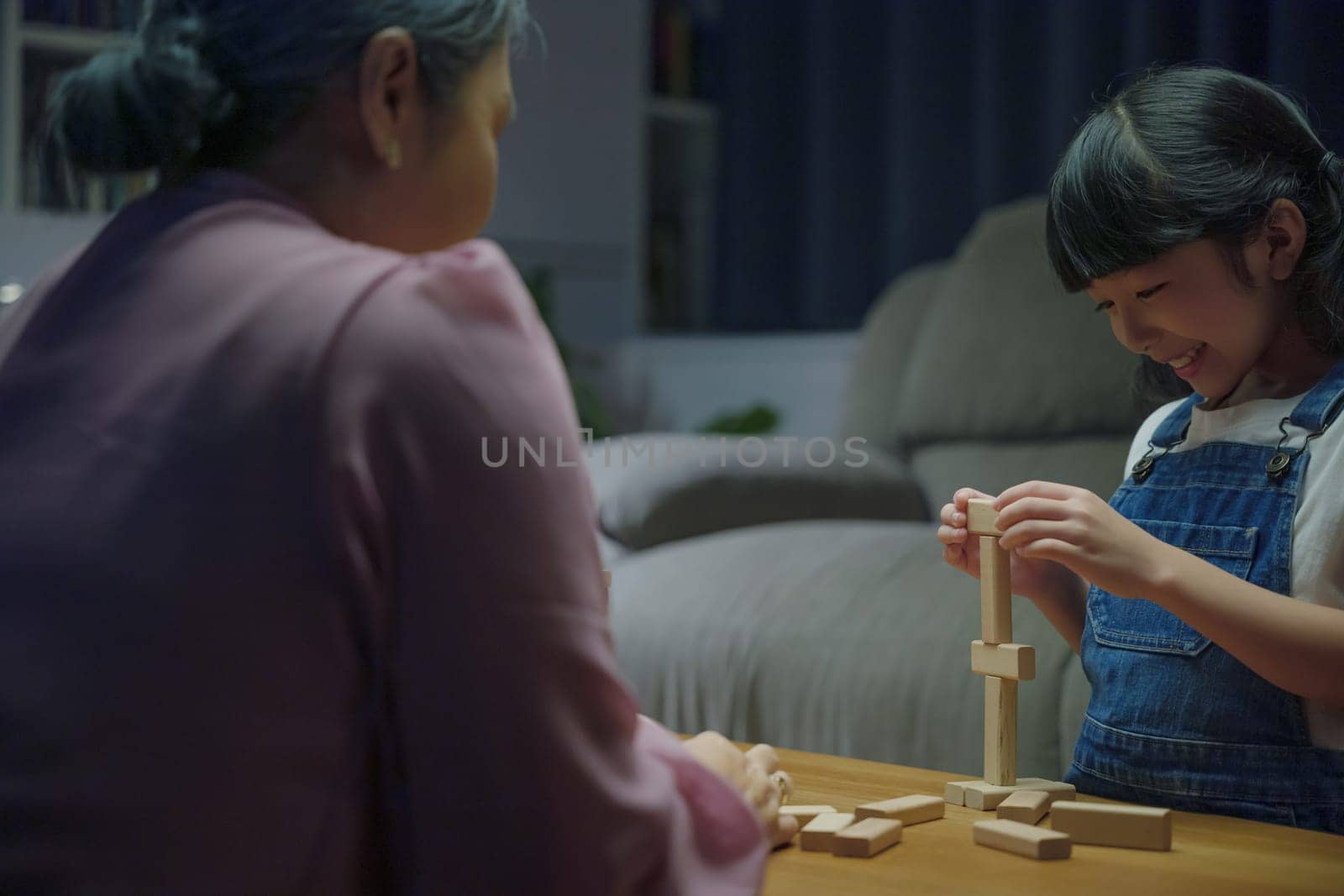 Smiling parent having fun play build constructor tower of wooden blocks, Happy grandparents Asian family enjoy playing toy block with little daughter together in home living room at night time