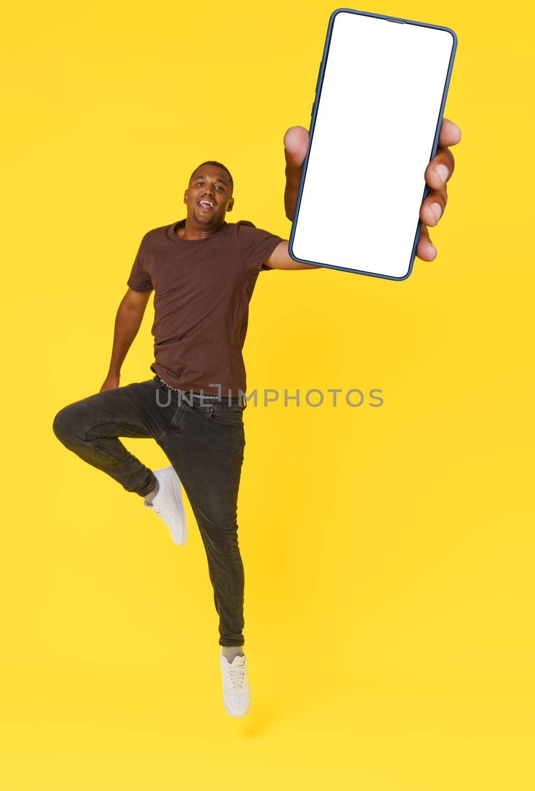 Energetic African American student is captured jumping with mobile phone featuring empty white screen in hand, on yellow background. This image is perfect for product placement or advertising concepts. by LipikStockMedia