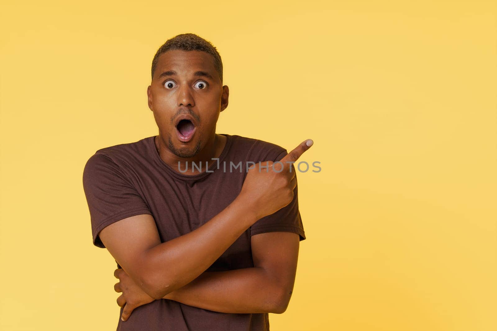 Amazed African American man is shown pointing to an empty copy space on a yellow background. This image is perfect for product placement or advertising concepts. High quality photo