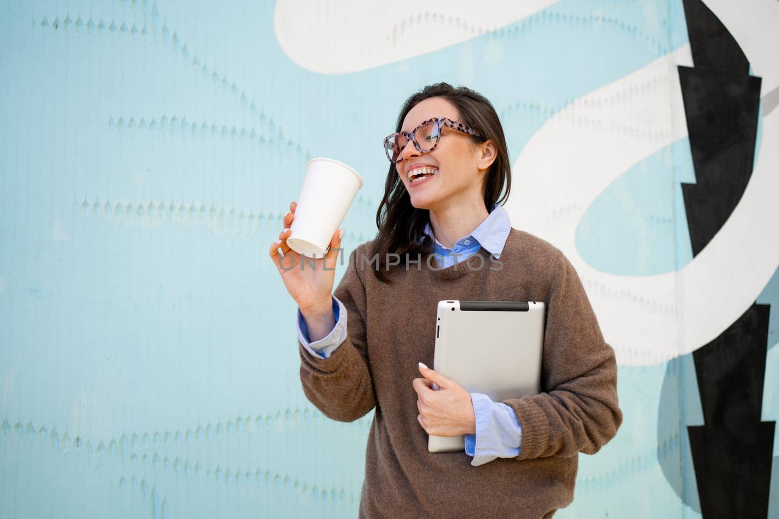 Positive business woman wearing glasses with leopard print and sweater walking down city street, holding coffee in paper cup mug and tablet computer