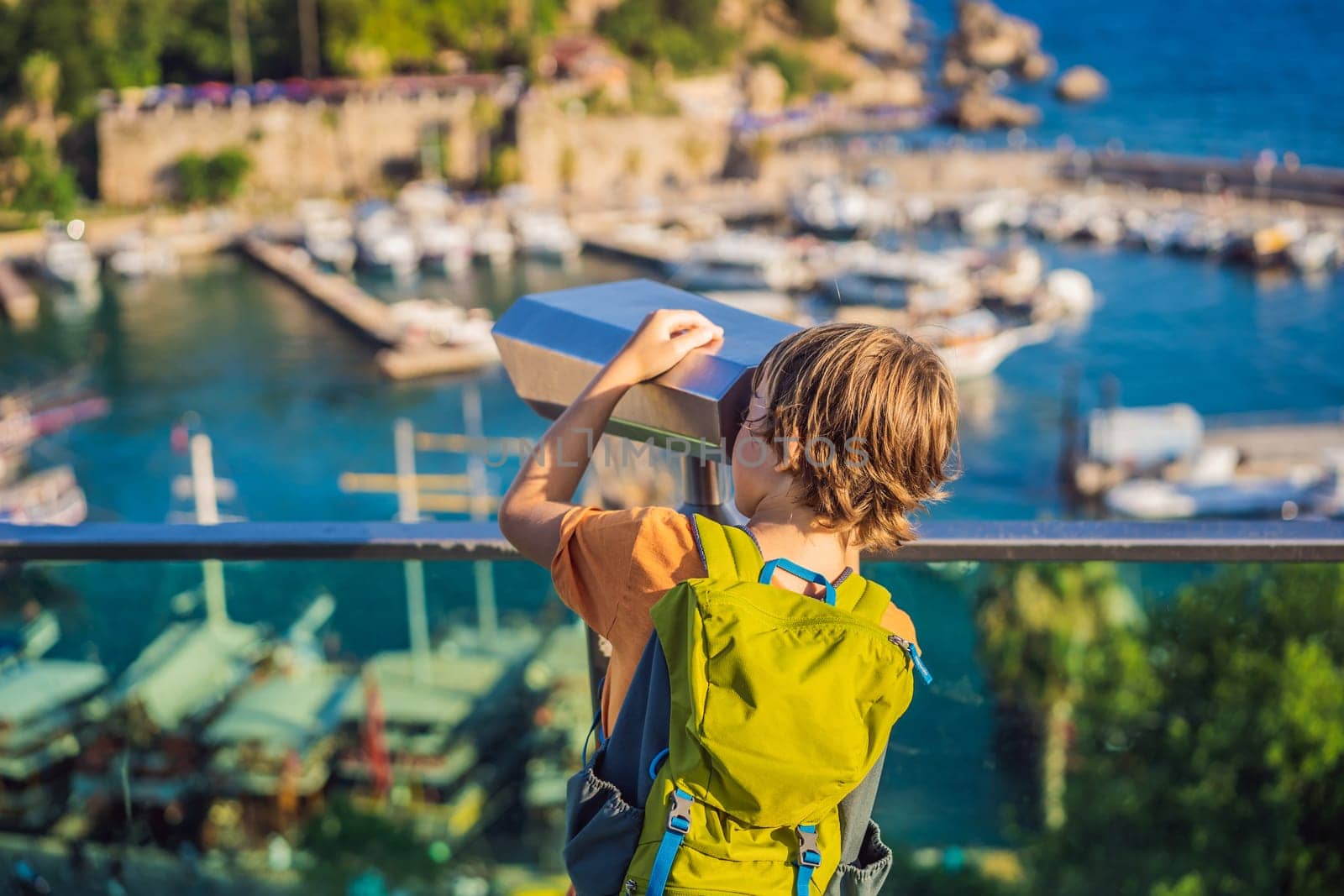 Boy tourist looks through binoculars in Old town Kaleici in Antalya. Turkiye. Panoramic view of Antalya Old Town port, Taurus mountains and Mediterrranean Sea, Turkey. Traveling with kids concept.
