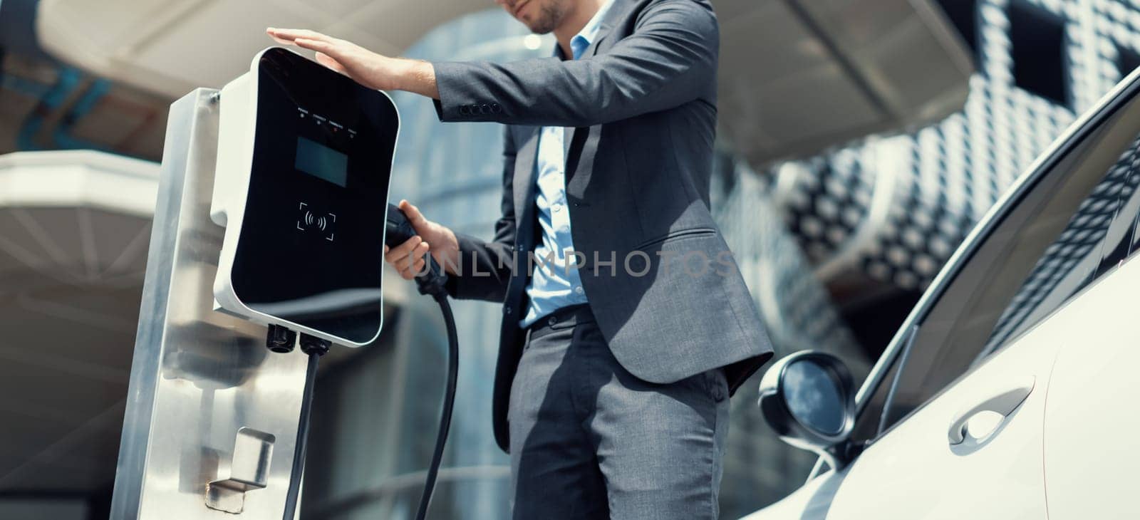 Below view of progressive businessman with electric car recharging at public charging station at modern city residential building. Eco friendly rechargeable EV car powered by alternative clean energy.