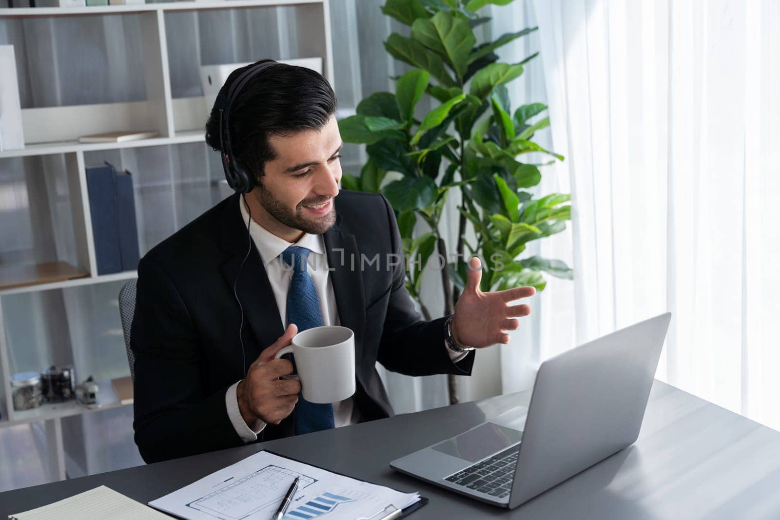 Manager of call center operator office sitting on his desk with his coffee while working on laptop. Office worker wearing headset and black suit working on customer support or telemarketing. fervent