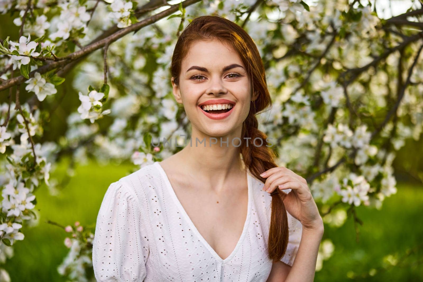 portrait of a joyful woman in a light dress against the background of a flowering tree, holding herself by her braid. High quality photo