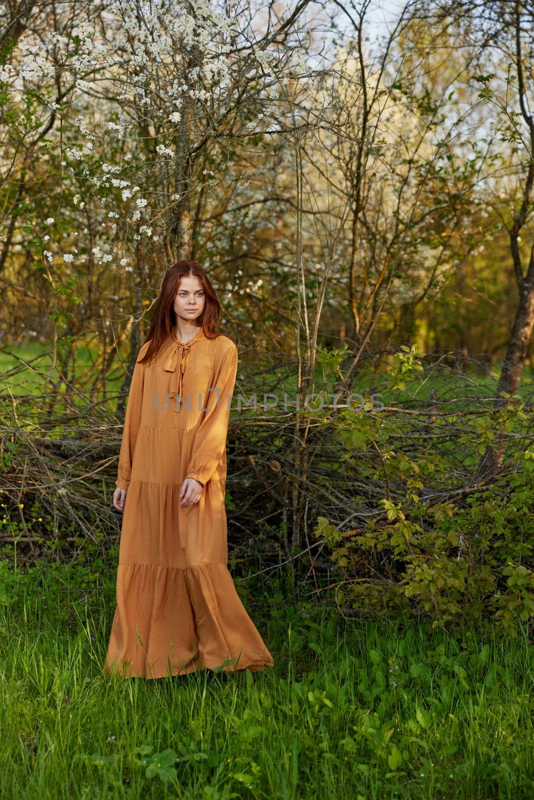 an elegant, sophisticated woman poses relaxed standing near a wicker fence at the dacha in a long orange dress. High quality photo