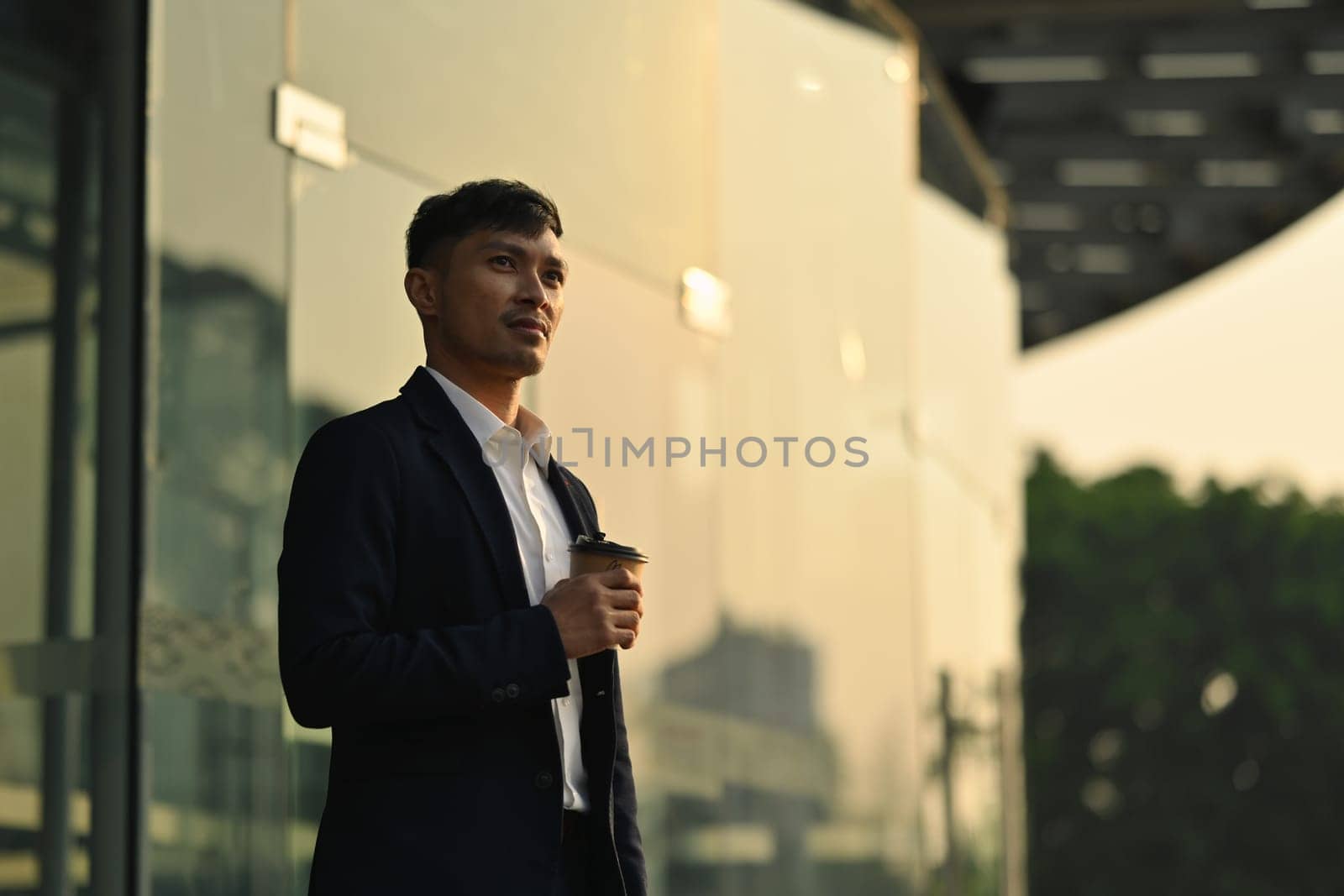 Thoughtful young businessman in formalwear standing outside building with cityscape background in early morning.