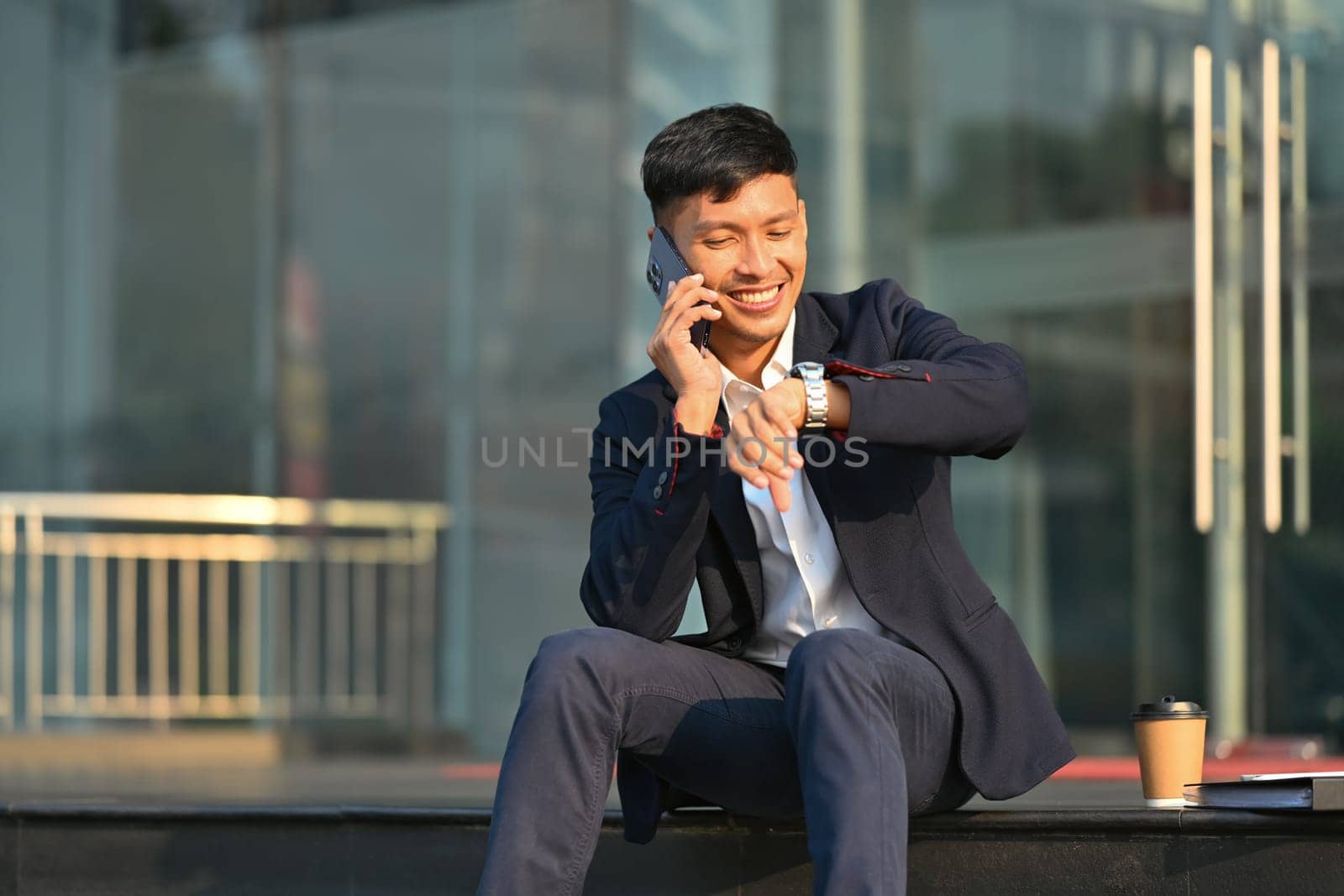 Smiling young businesswoman in formal clothes talking on mobile phone and checking time on wristwatch.