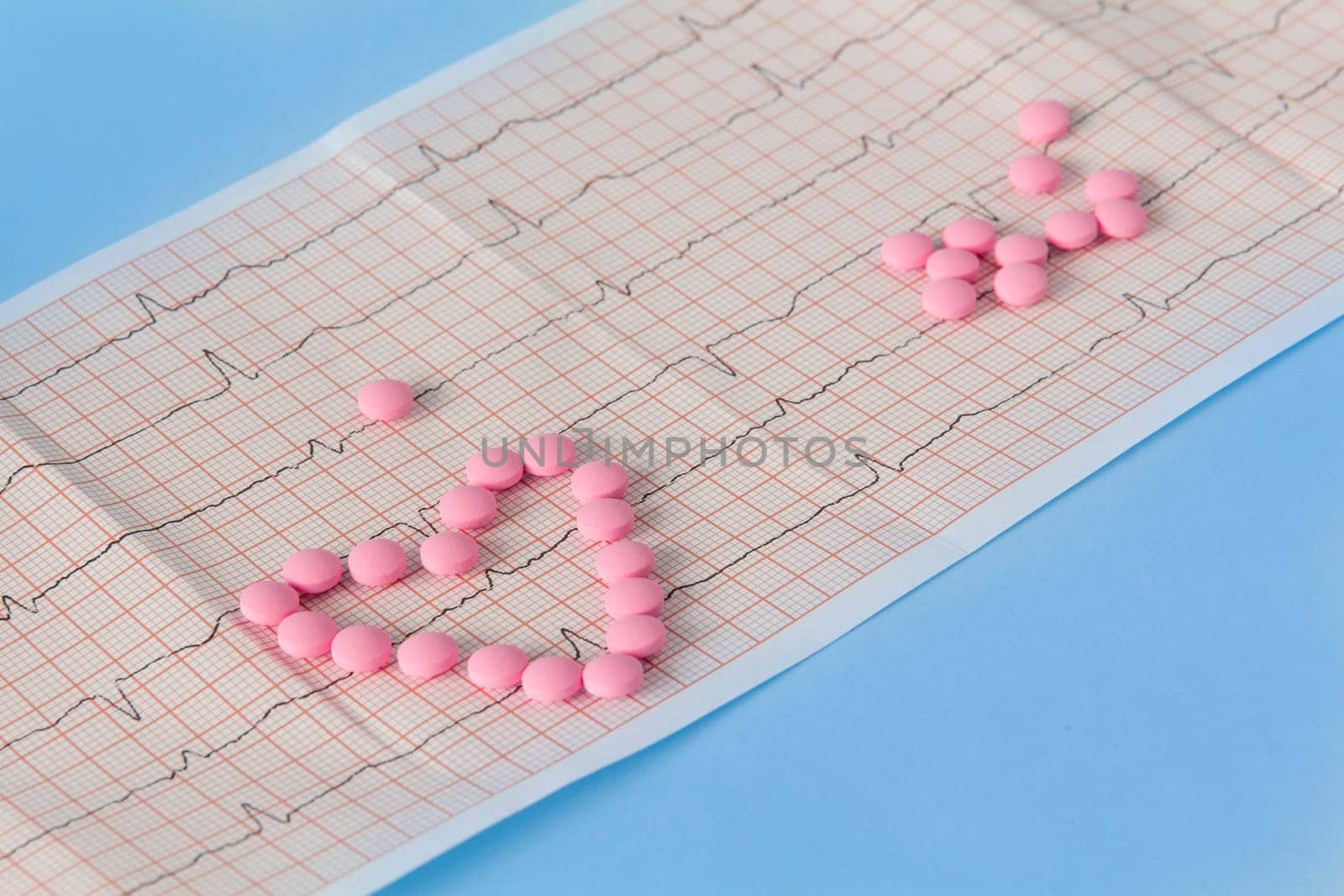 A large handful of pink pills lie in the form of a heart on an electrocardiogram, on a blue background. The concept of a healthy lifestyle and timely medical examination. by Alla_Yurtayeva