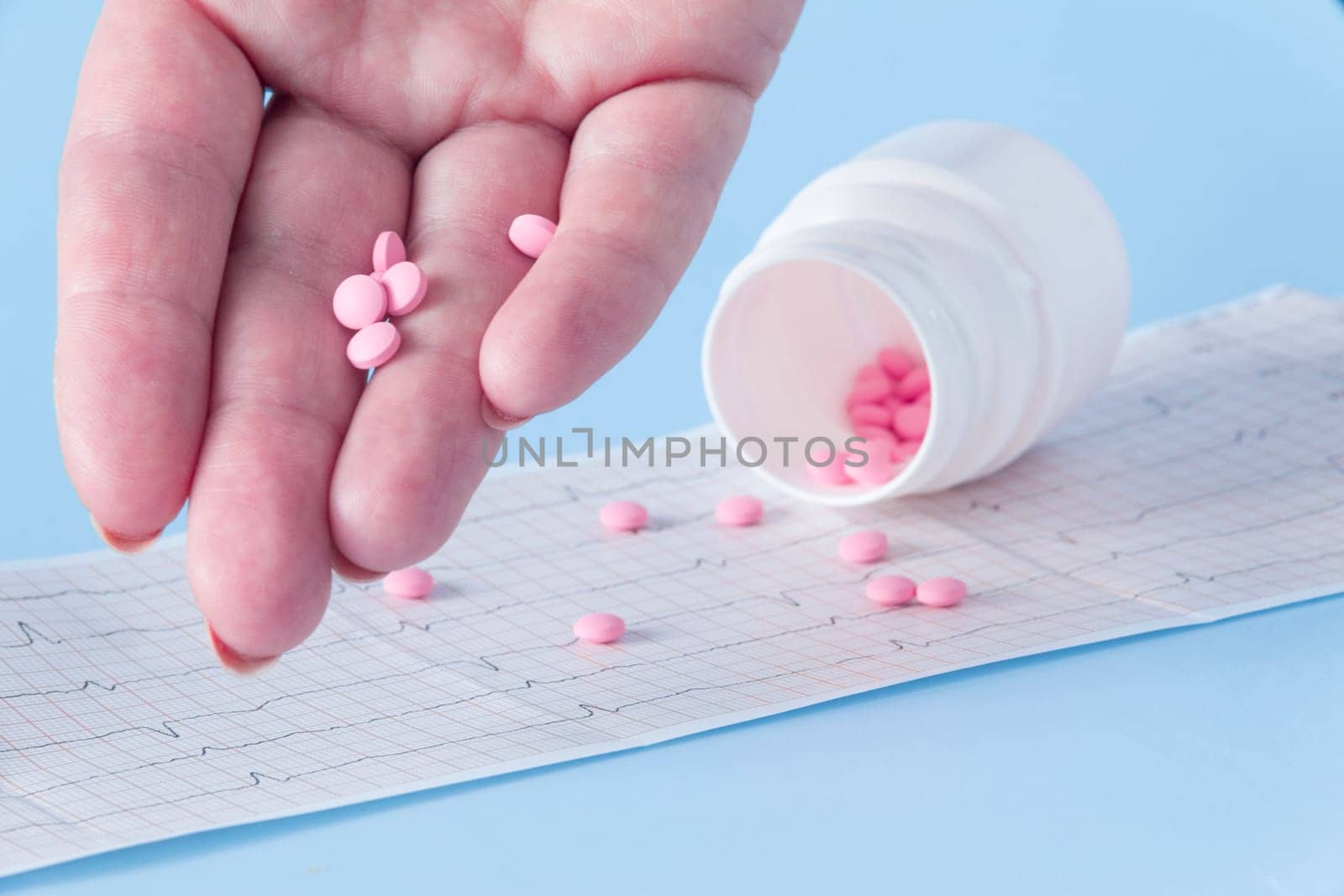 A hand pours a handful of pink pills from a white jar onto an electrocardiogram of the heart, on a blue background. The concept of a healthy lifestyle and timely medical examination