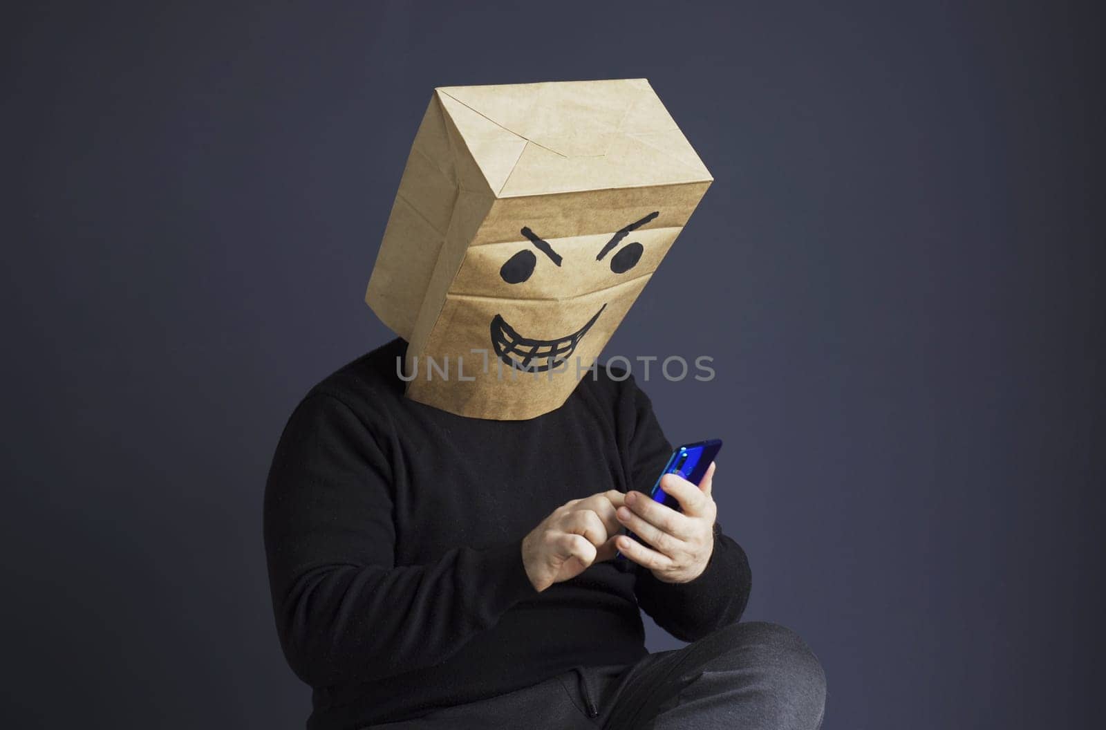 A man in a black turtleneck with a paper bag with an angry emoji on his head is typing a message on his smartphone while sitting on a chair. Emotions and anger