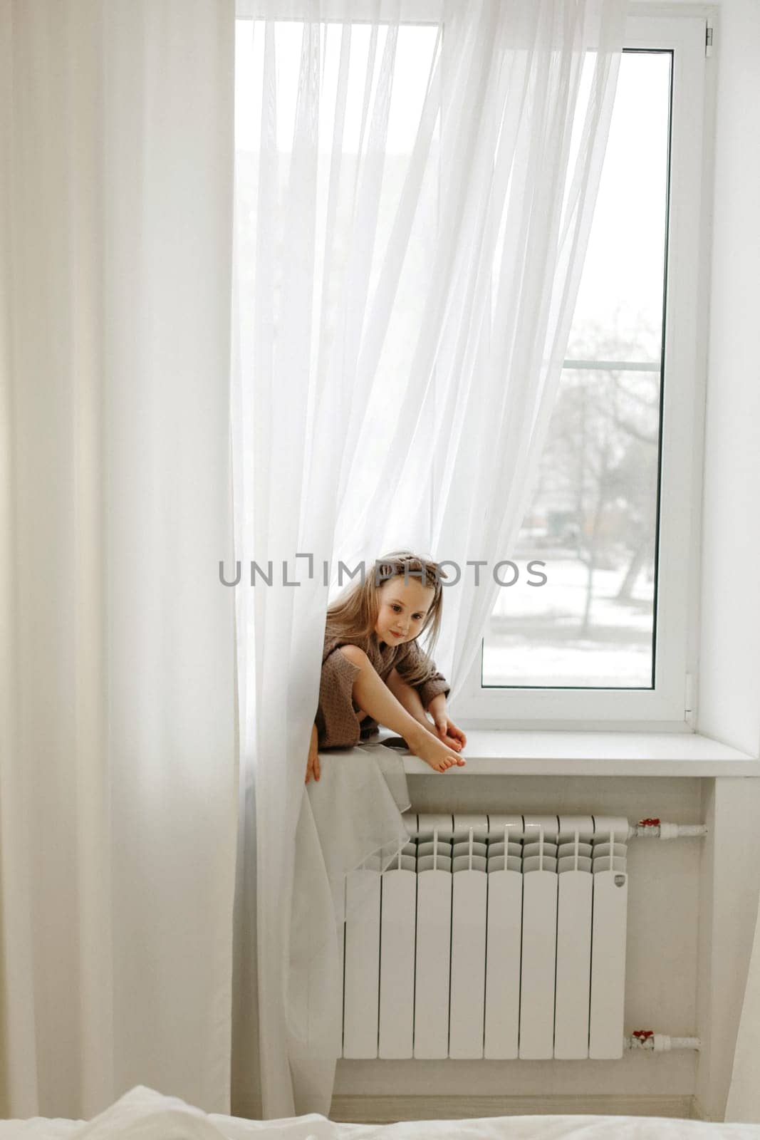 Portrait of a girl in a brown dressing gown, who sits on the windowsill and examines her toes.