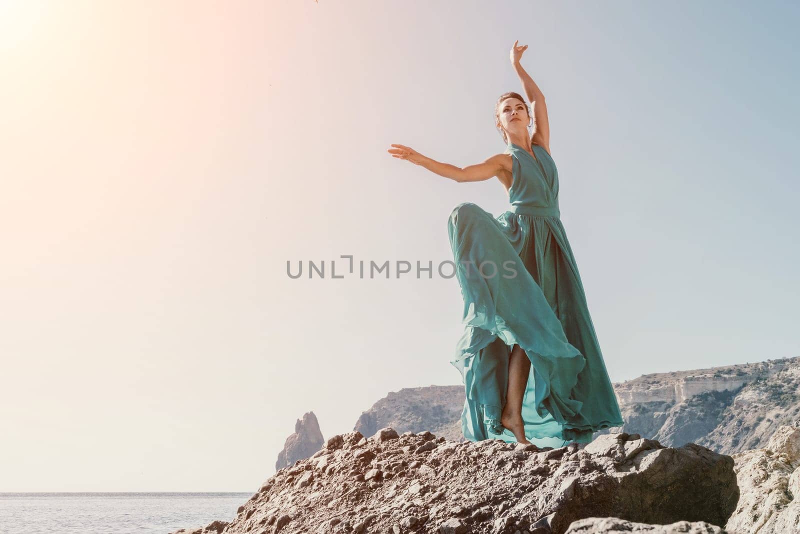 Side view a Young beautiful sensual woman in a red long dress posing on a rock high above the sea during sunrise. Girl on the nature on blue sky background. Fashion photo.