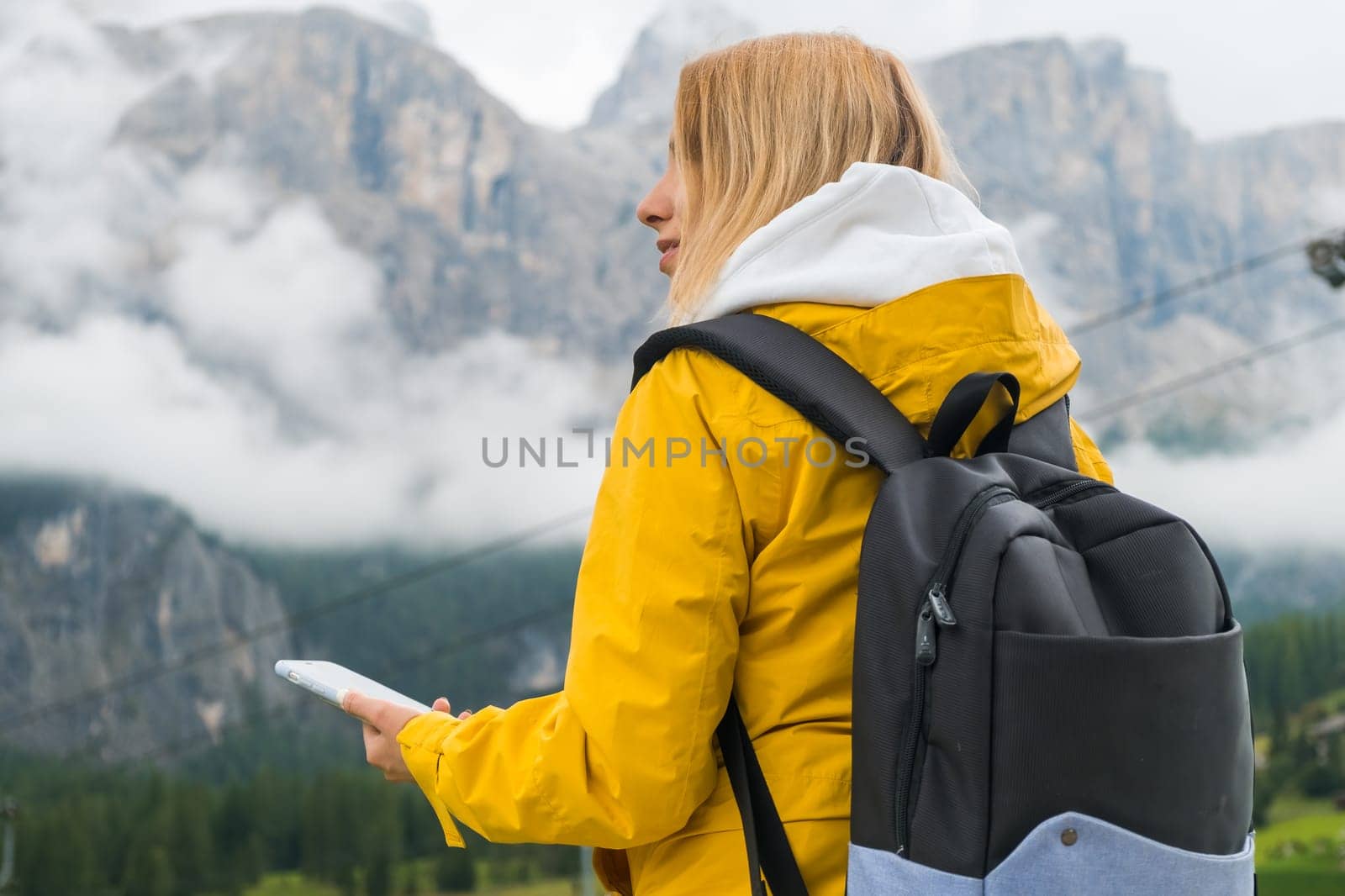 A blonde hair woman in a yellow sport jacket and blue backpack checks the route using a smartphone in Dolomites Alps