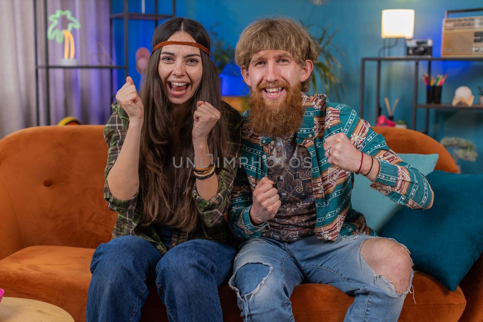 Young family married hippie couple man woman shouting, celebrating success, winning, wow, goal achievement, good news, lottery jackpot luck at home. Husband and wife together watching TV on room sofa