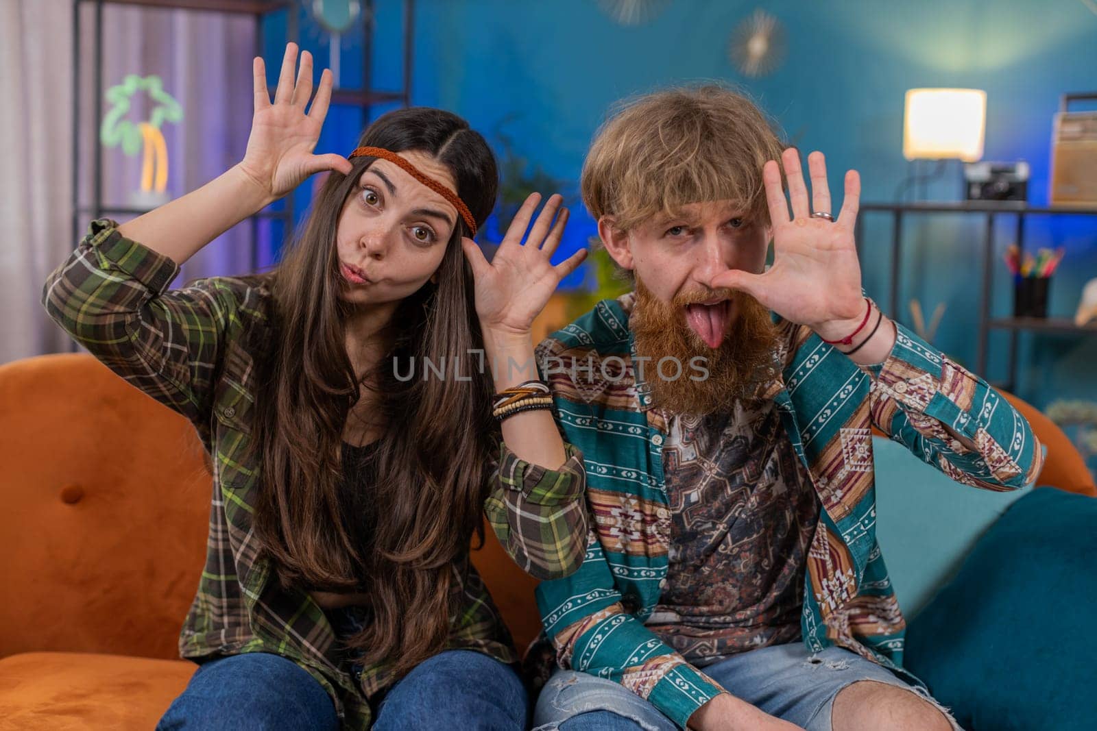 Family marriage hippie couple man woman making playful silly facial expressions and grimacing fooling around showing tongue. Husband and wife together on sofa in room. Domestic life leisure activities