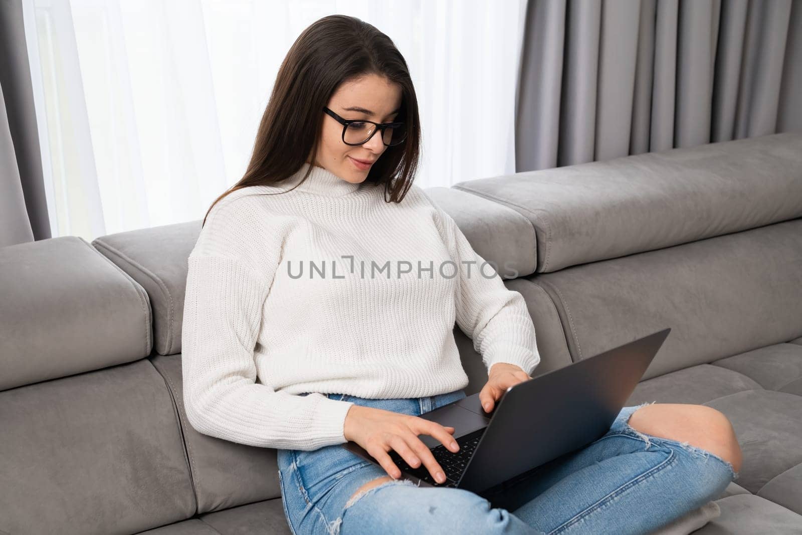 Female student gets happy to see good final results of examination on Notebook. Young woman sits on sofa with laptop in living room feeling joy