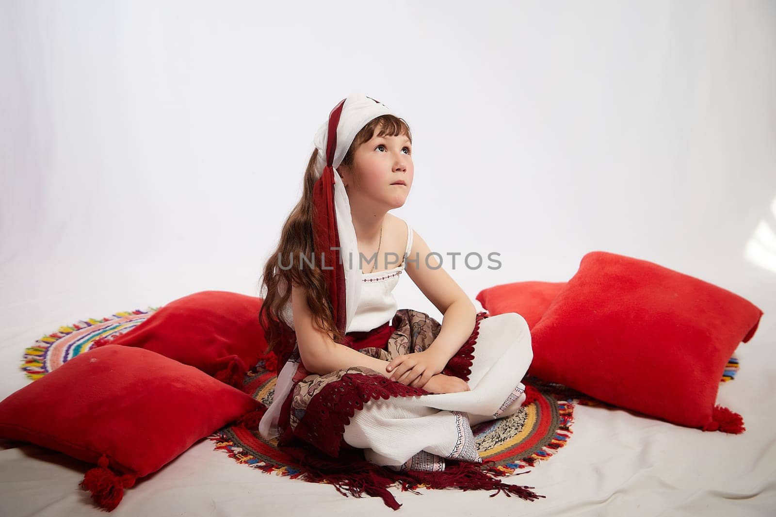 Portrait of Little girl in a stylized Tatar national costume having rest with a red pillow on a white background in the studio. Photo shoot of funny young teenager who is not a professional model