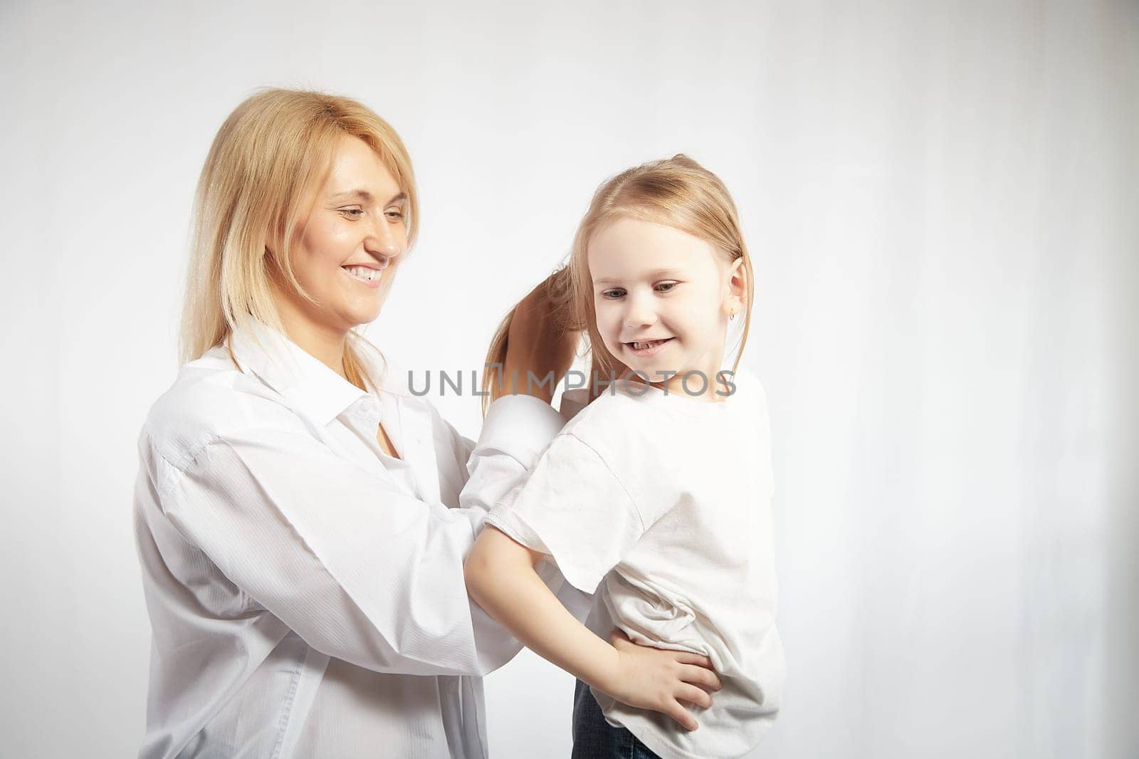 Close portrait of blonde mother and daughter where mom braids hair and makes ponytail and hairstyle for her girl on white background in studio. The concept of love, friendship, caring in family