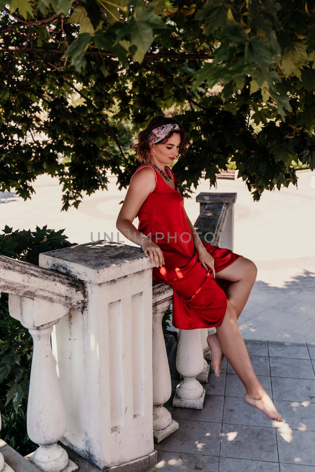 woman in a red silk dress and a bandage on her head smiles against the background of the leaves of a tree. She is leaning on the coop and looking into the camera. Vertical photo