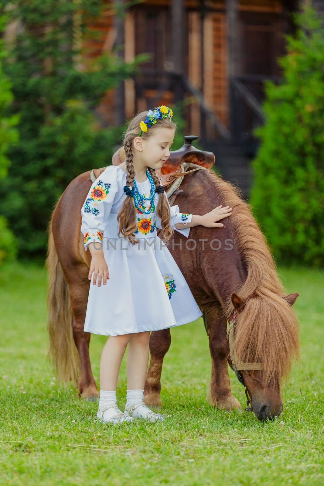 young girl in Ukrainian national dress strokes a pony that grazes on the lawn