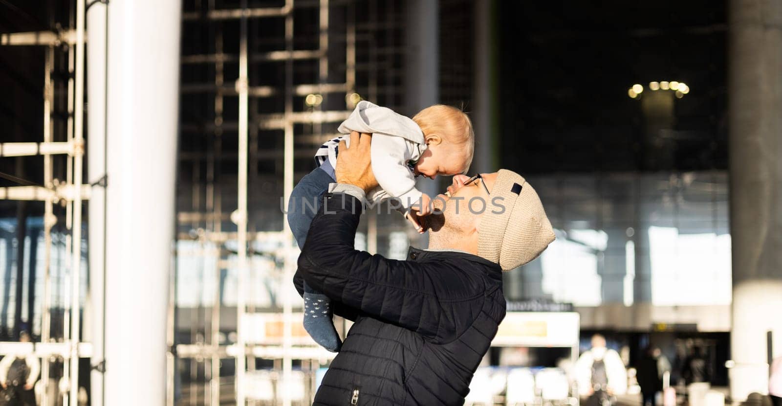 Father happily holding and lifting his infant baby boy child in the air after being rejunited in front of airport terminal station. Baby travel concept