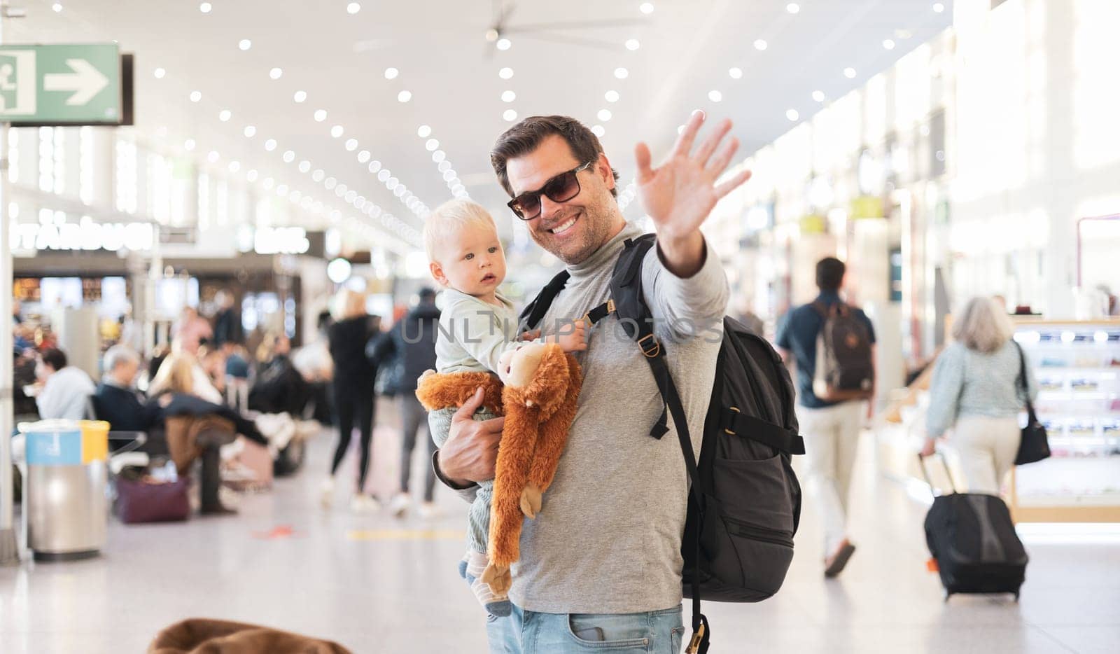 Father traveling with child, holding his infant baby boy at airport terminal waiting to board a plane waving goodby. Travel with kids concept. by kasto